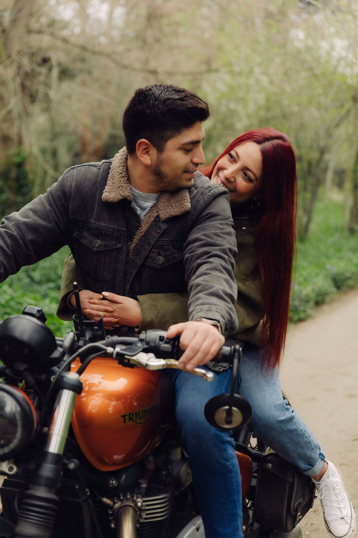 A couple sits together on an orange Triumph motorcycle on a tree-lined path, sharing a tender moment.
