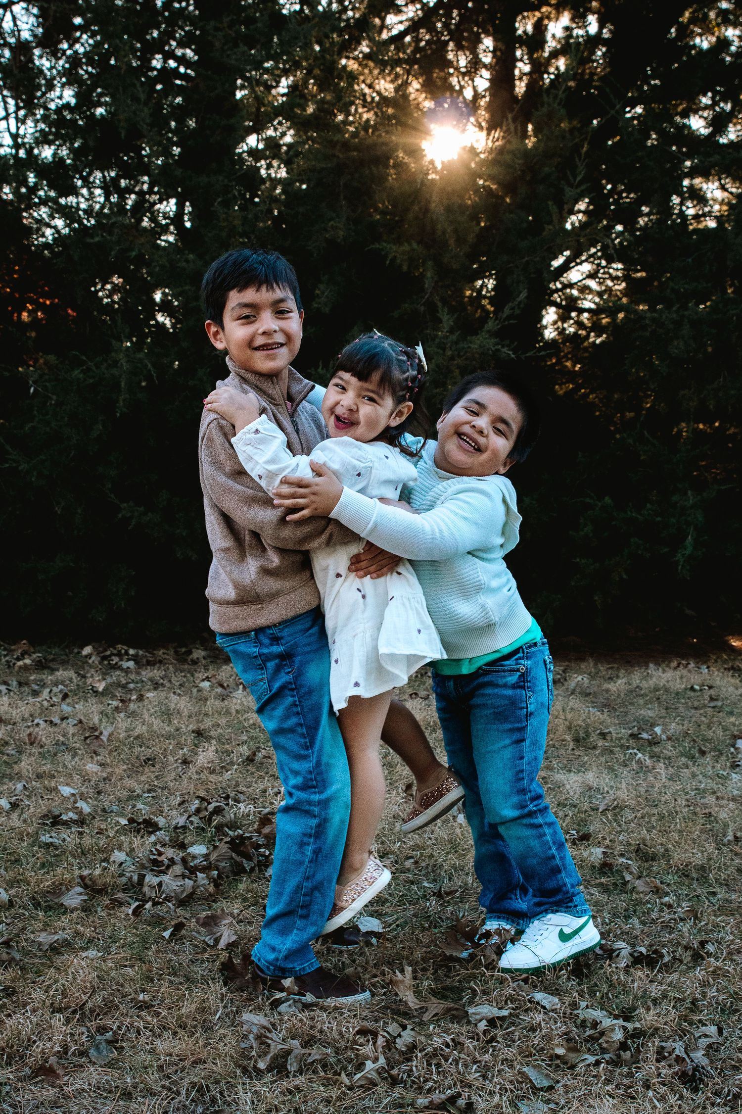Three happy children hugging and laughing outdoors at sunset with trees in the background.