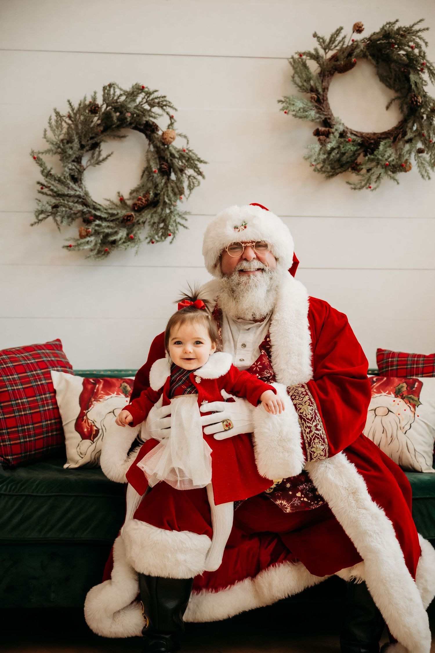 Baby girl in red dress sits on Santa's lap in front of festive wreaths and holiday pillows.