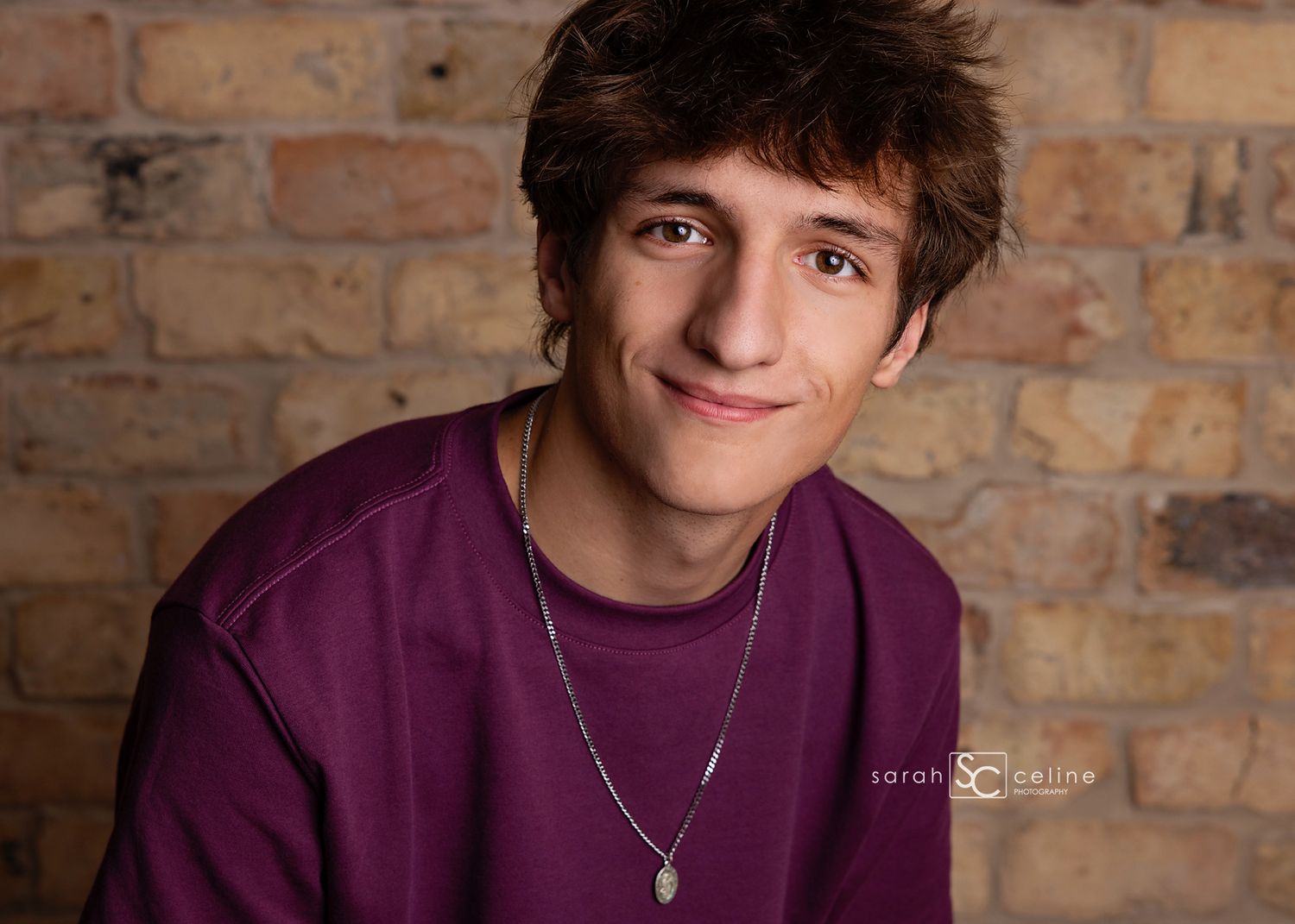 Smiling teenage boy in purple shirt with necklace poses for senior portrait against rustic brick wall.