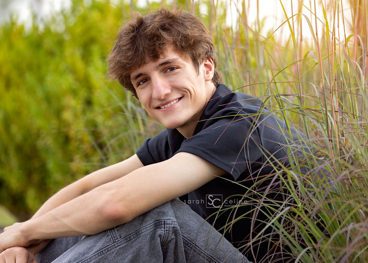 A smiling young man with curly hair sits outdoors among tall green grasses in a casual black shirt.
