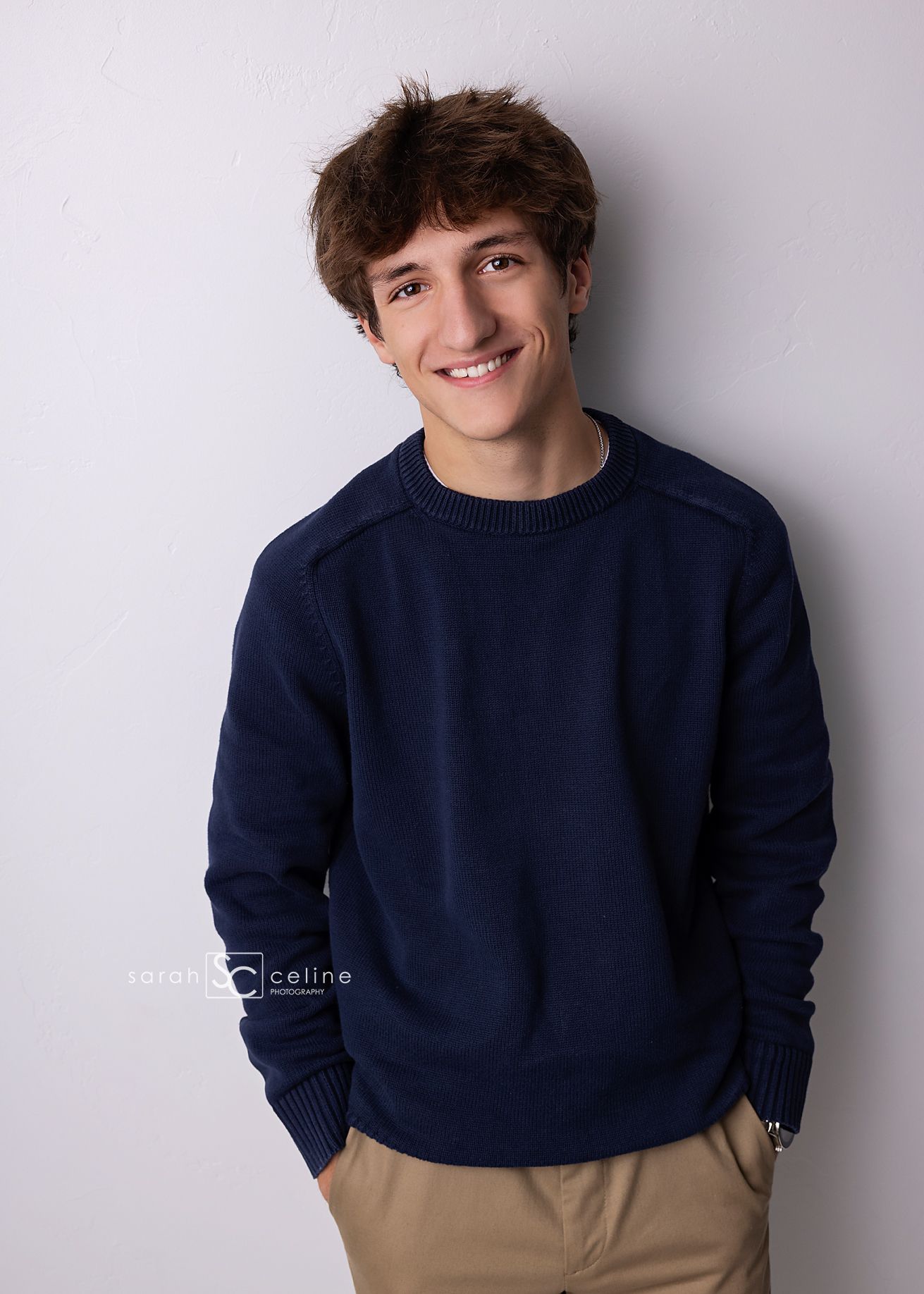 Young man in navy sweater and khaki pants smiling confidently against white background in senior portrait photo.