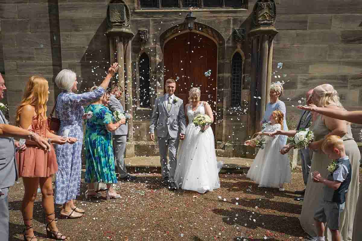 Bride and groom exit church doors as guests throw colorful confetti during outdoor wedding celebration.