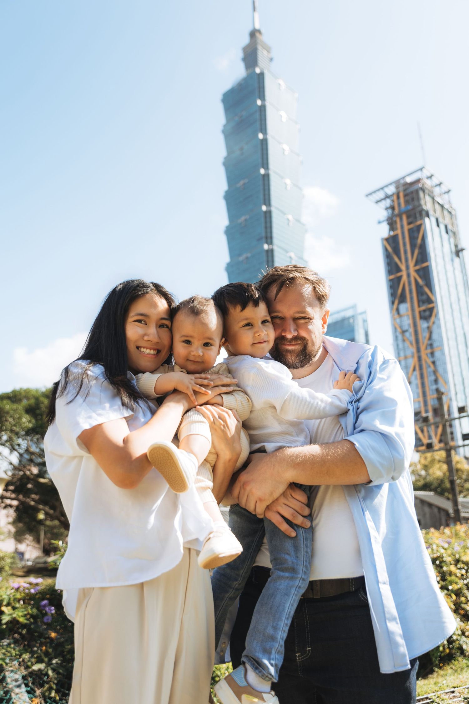 Happy family with two young children posing outdoors with Taipei 101 skyscraper in the background on a sunny day.