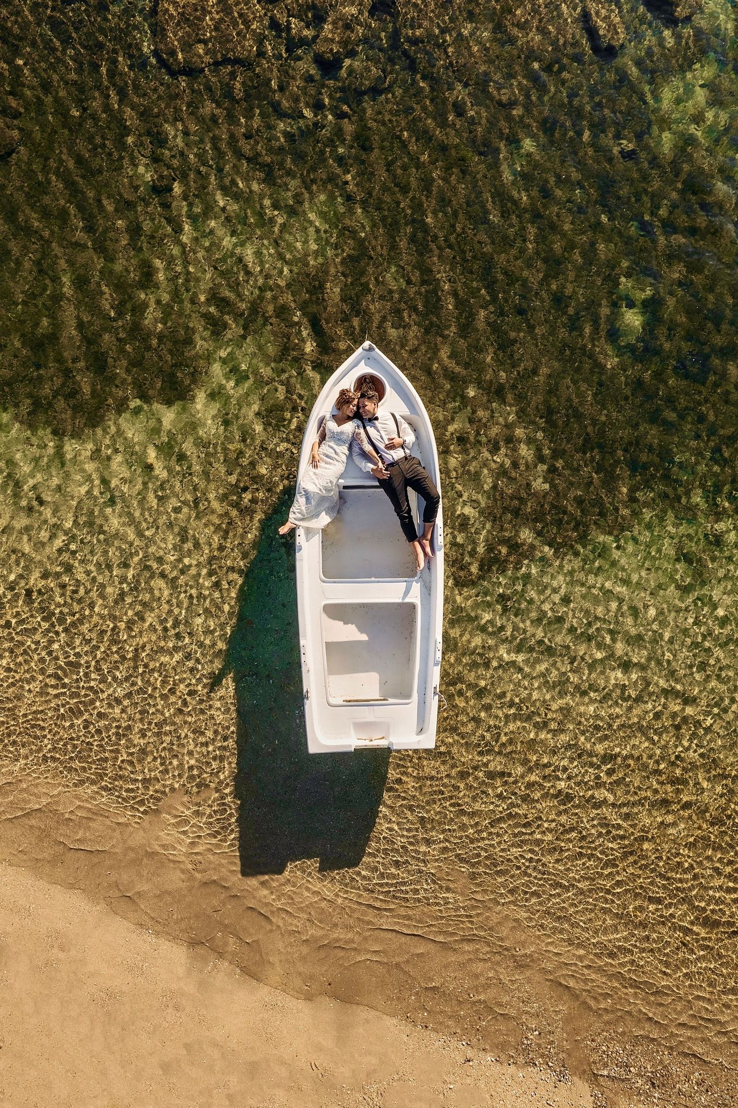 Aerial view of a couple lying in a white boat near a sandy shore with clear shallow water.