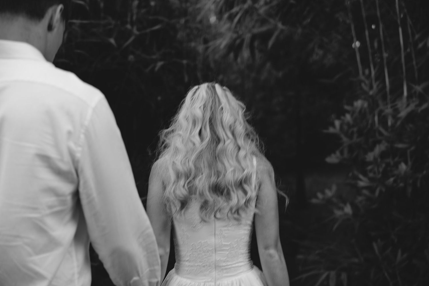 Black and white photo of a bride and groom walking hand in hand, viewed from behind, surrounded by lush greenery.