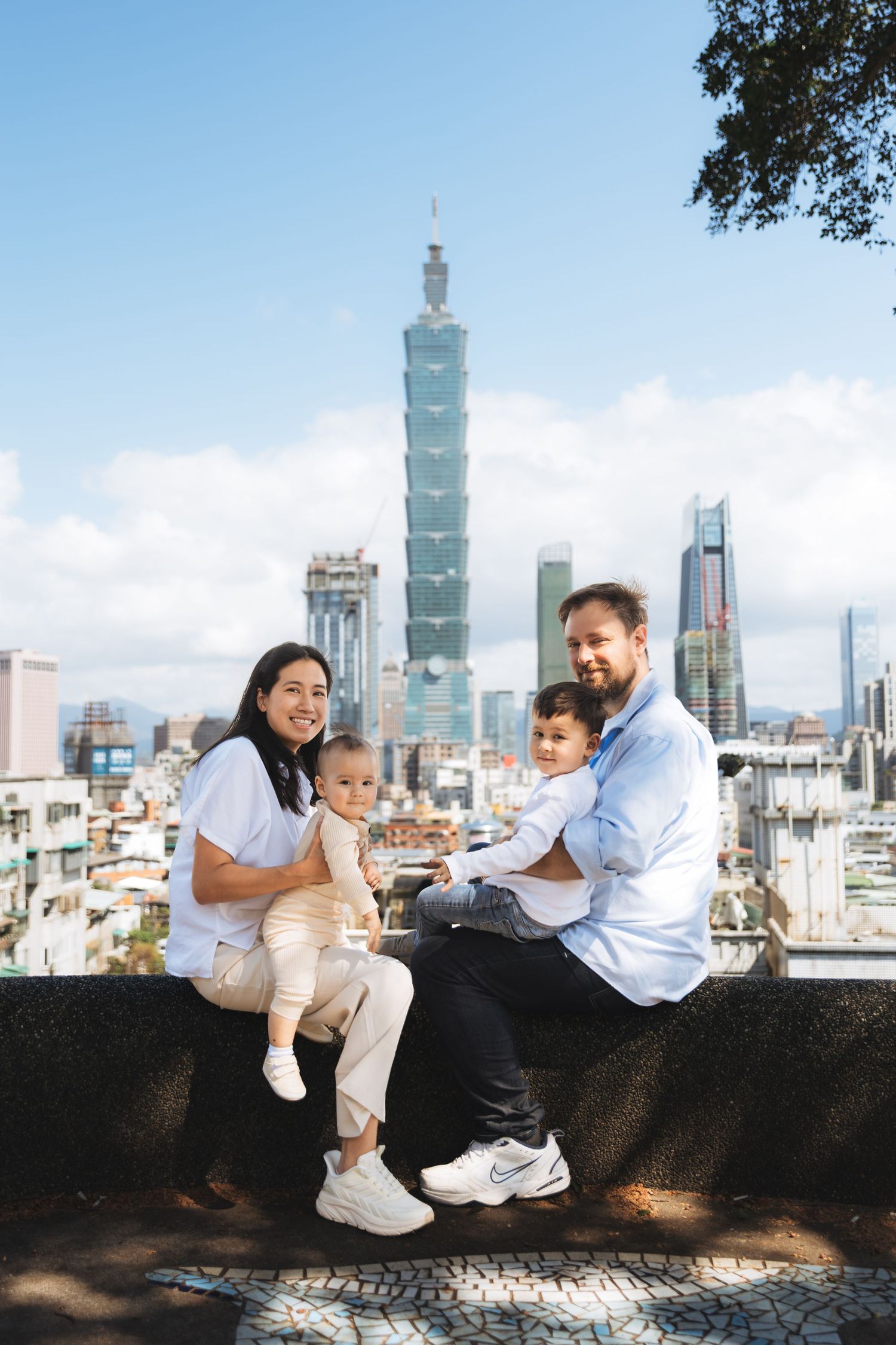 Family of four posing with Taipei 101 skyscraper skyline backdrop on a sunny day.