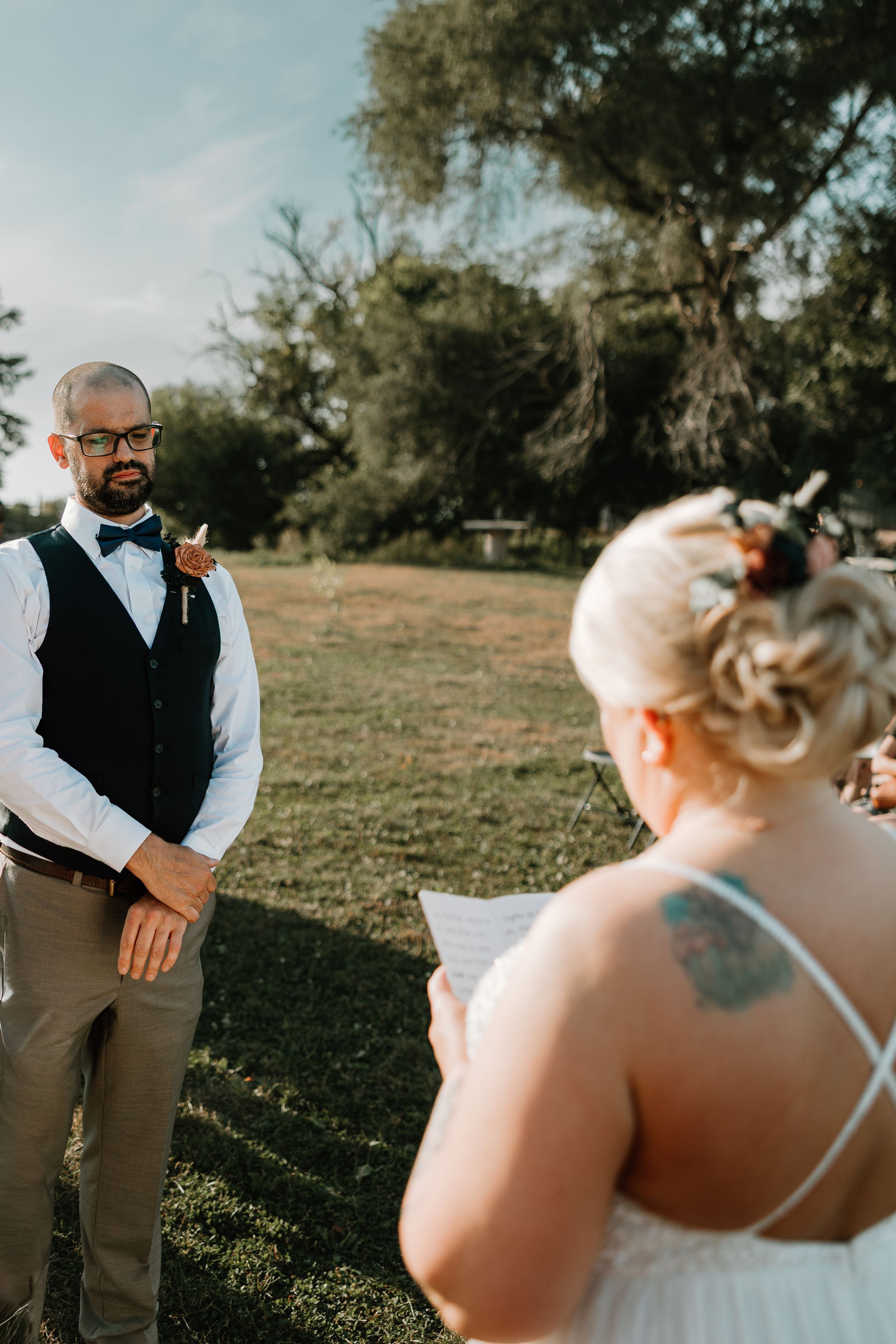 Groom in vest and bow tie listens emotionally as bride reads vows outdoors in golden light.