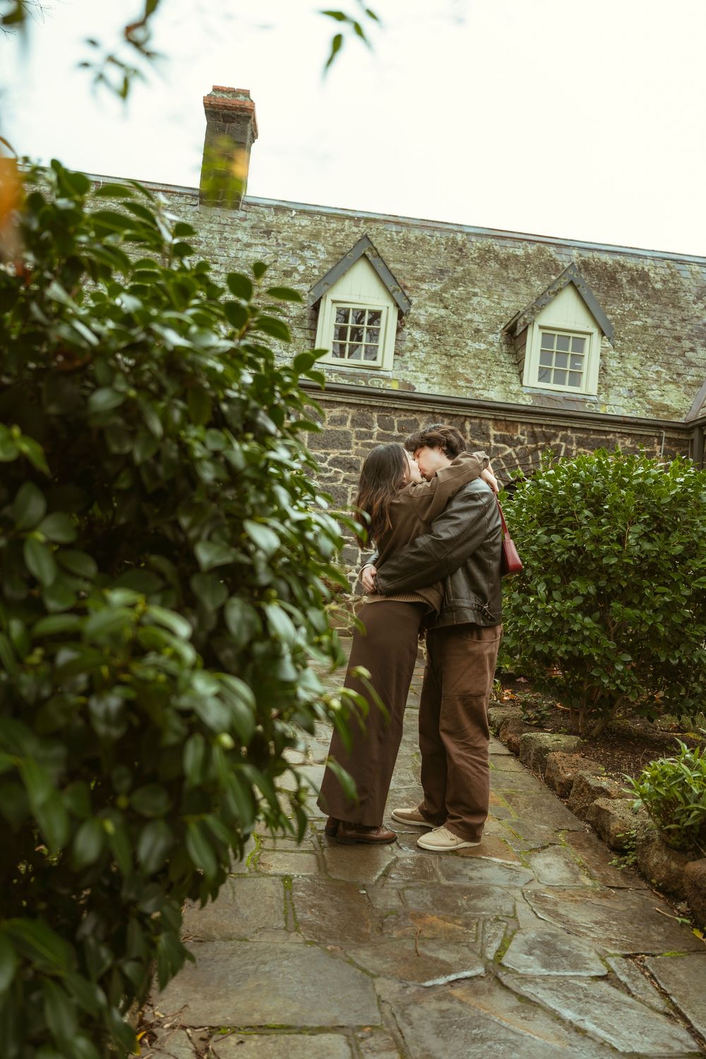 Couple embracing and kissing on a stone pathway beside a charming cottage with dormer windows and lush greenery.