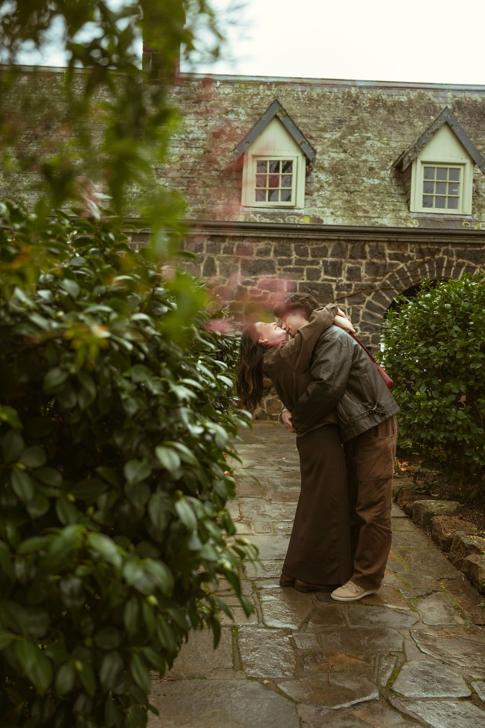 Couple sharing a romantic kiss in a lush garden pathway beside a charming stone cottage.