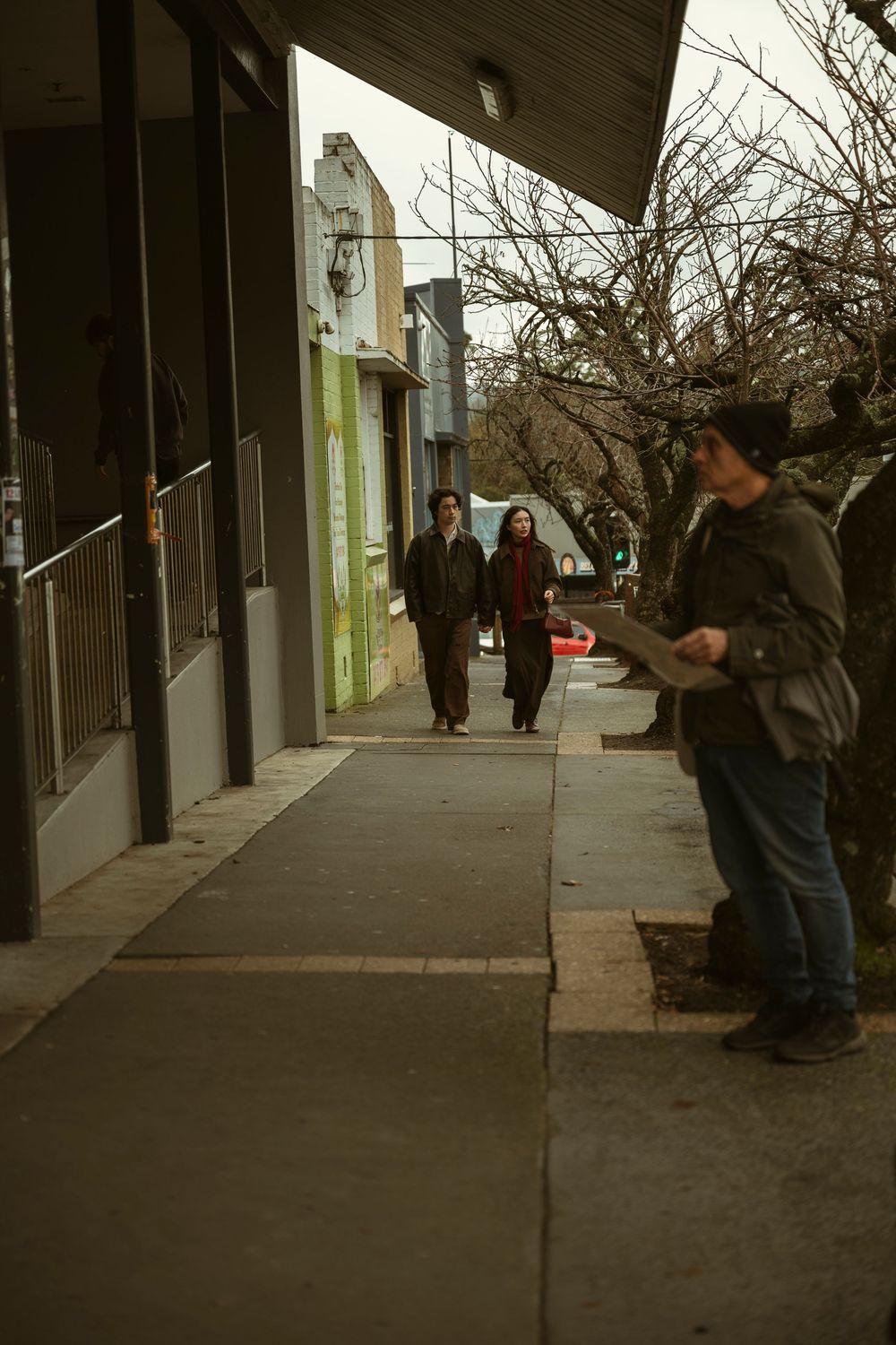 Three people walking on a city sidewalk past storefronts on a cloudy day, with bare trees lining the street.