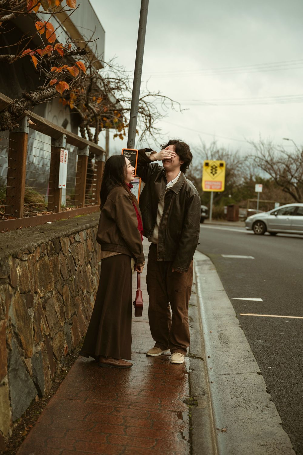 Couple standing on a wet sidewalk beside a stone wall on an overcast day, sharing a tender moment.