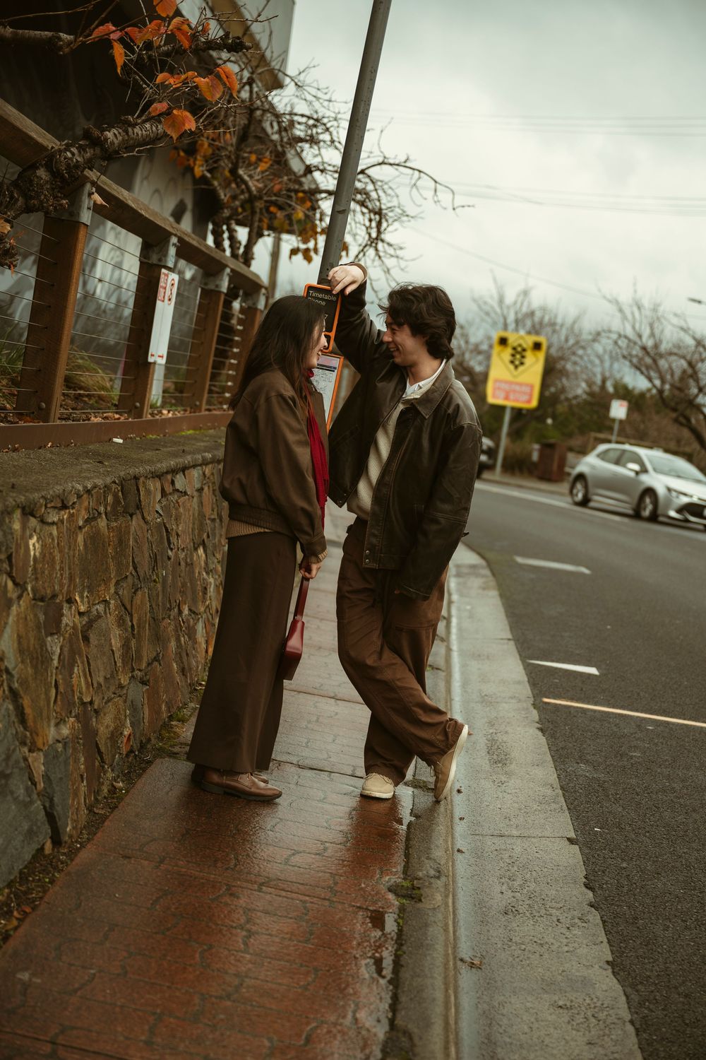 Couple sharing a romantic moment on a wet sidewalk beside a stone wall on a cloudy day.