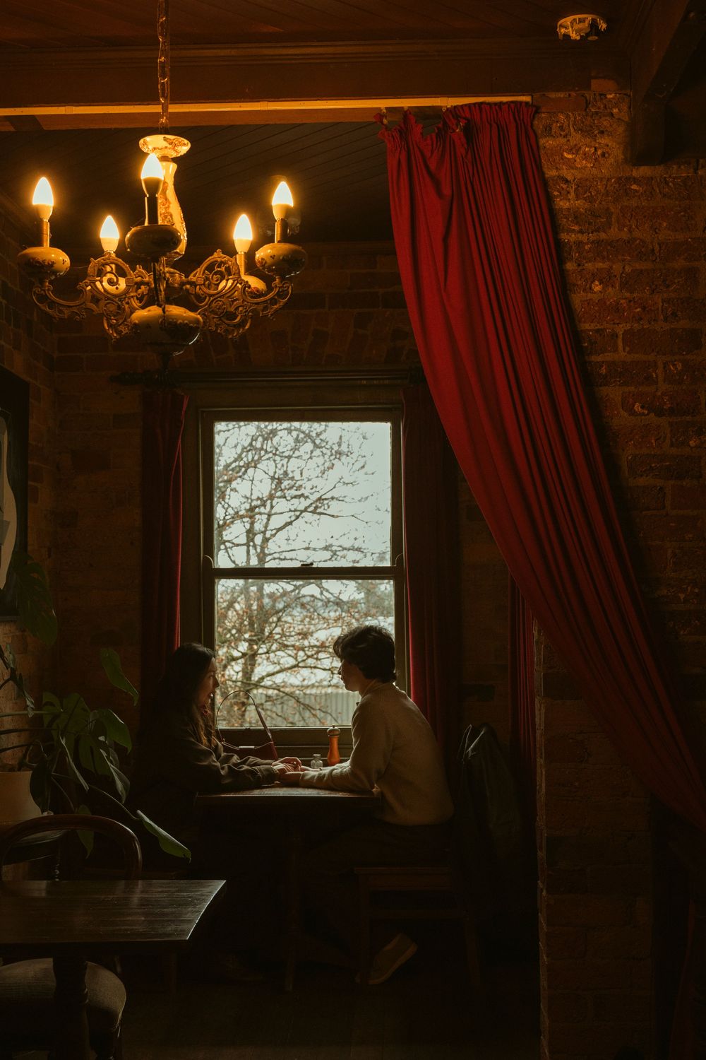 Two silhouetted figures dining by a window in a cozy rustic restaurant with a chandelier and red curtains.