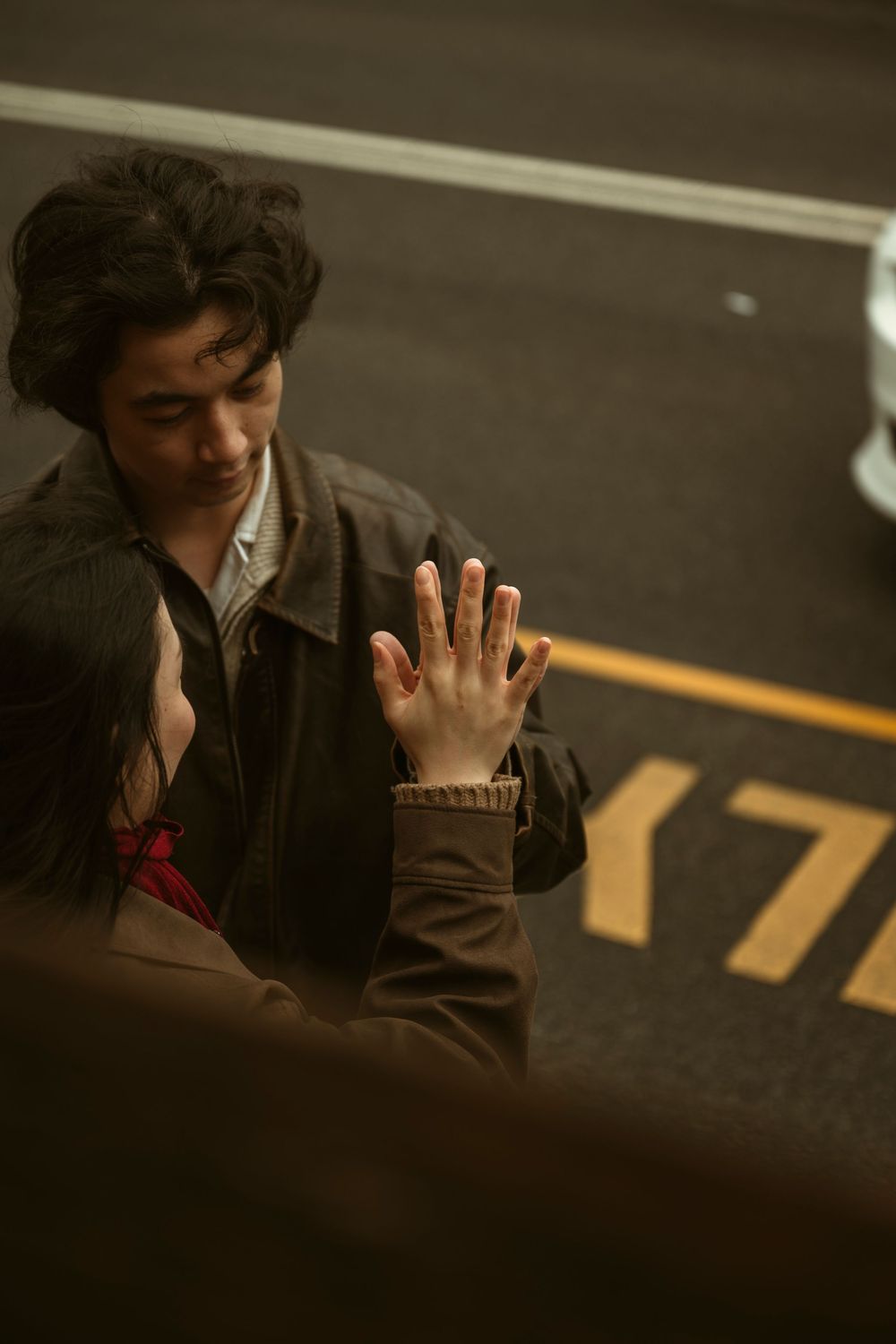 A young man in a brown jacket smiles as someone places their hand on his chest on a city street.