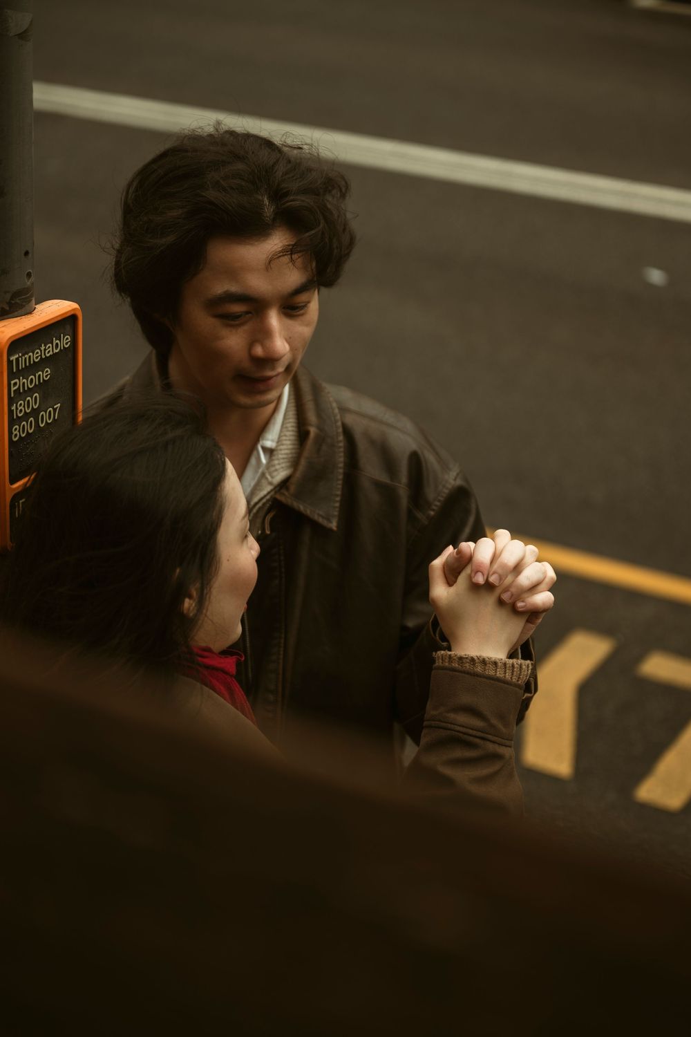 Two young Asian people holding hands at a bus stop at night, sharing a tender romantic moment.