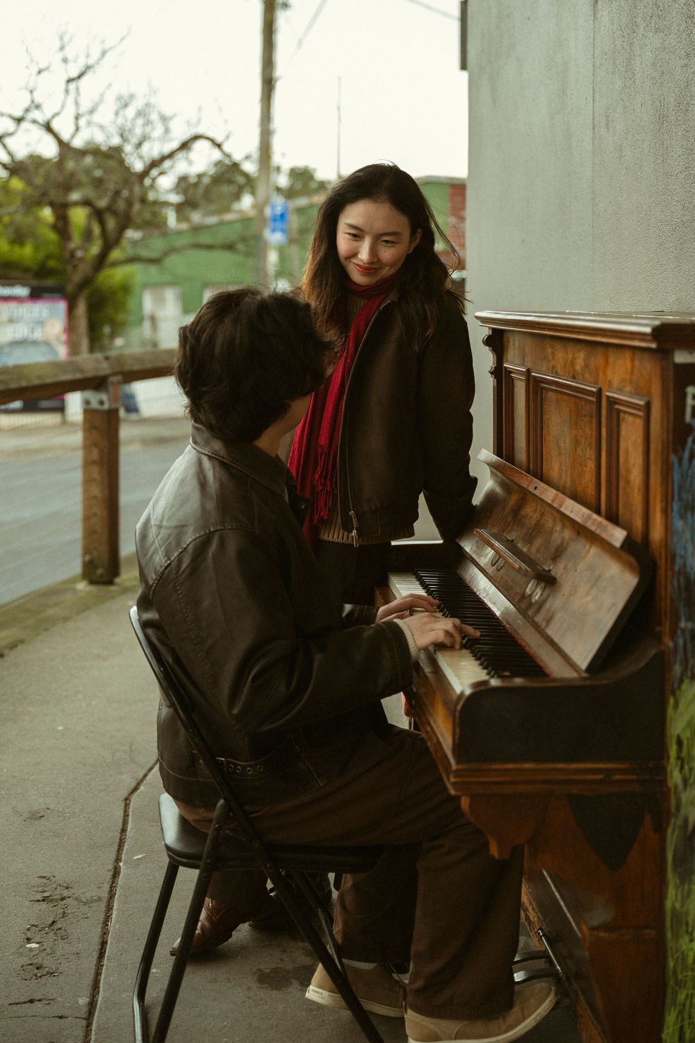A man plays an upright piano outdoors on a sidewalk while a smiling woman in a red scarf watches him.