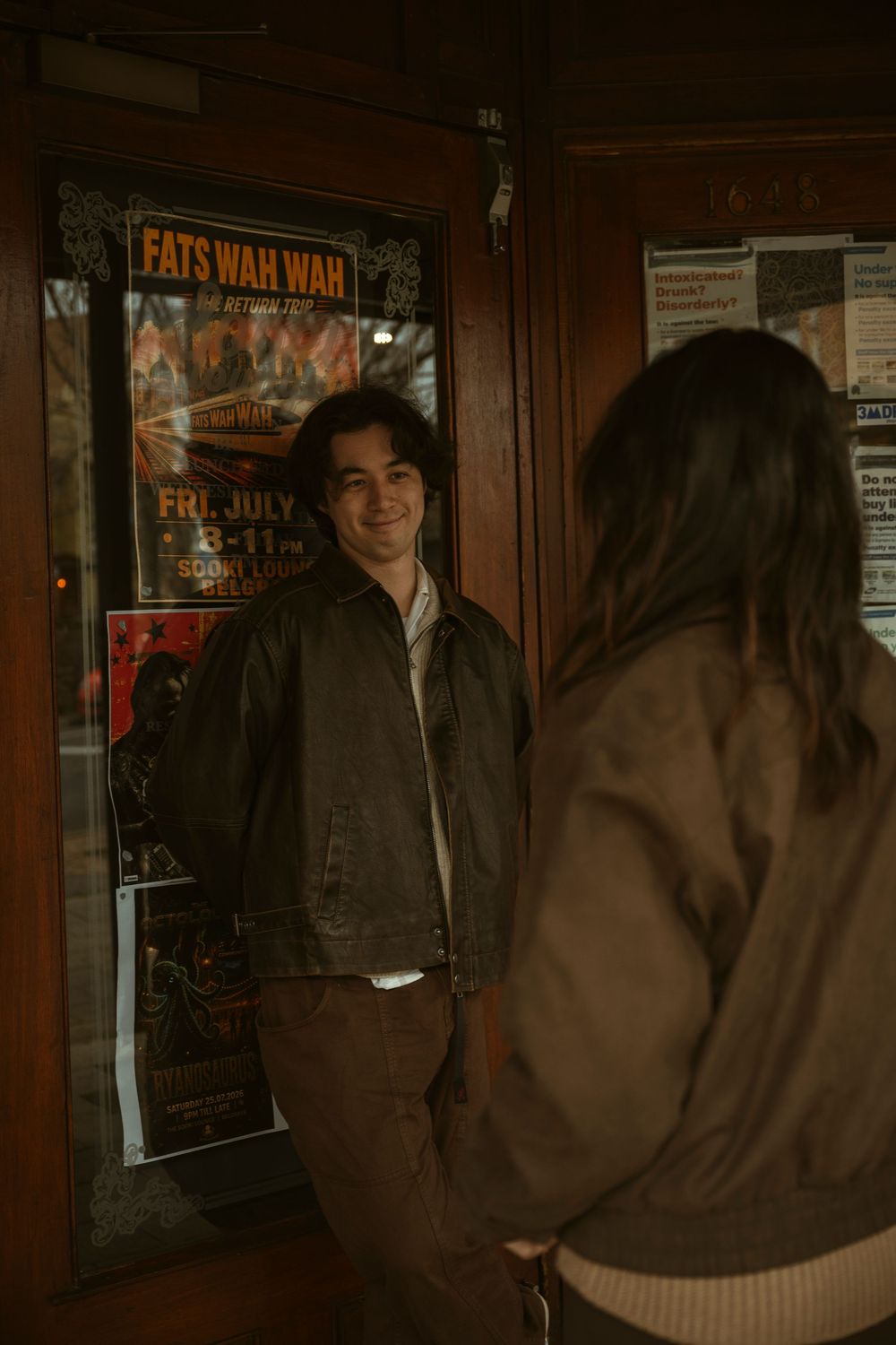 A smiling young man in a leather jacket leans against a door with concert posters.