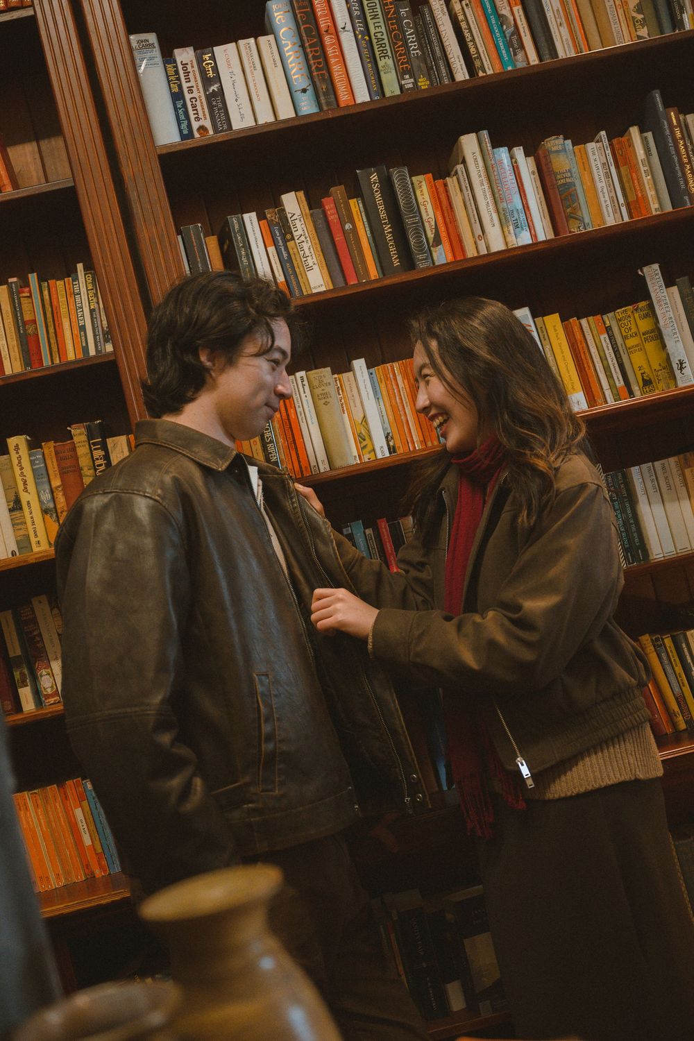Two people in leather jackets smile at each other in a cozy bookstore with wooden shelves full of colorful books.