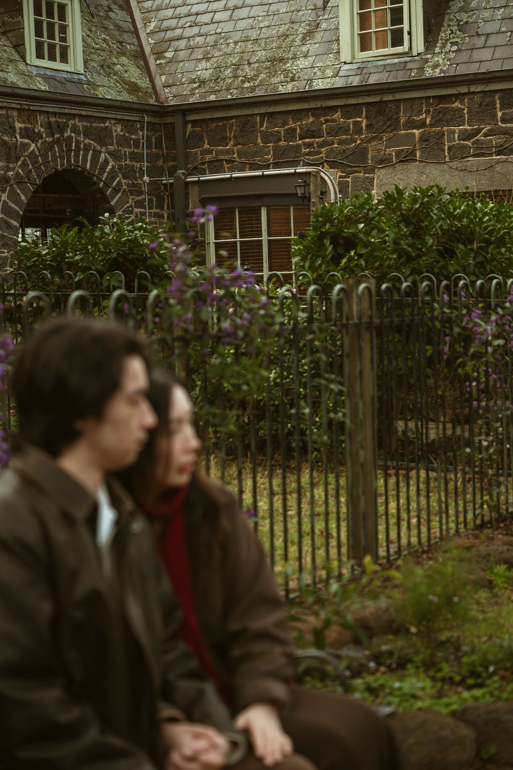 Couple sitting outdoors near vintage iron fence and stone building with lush green shrubs and purple flowers.
