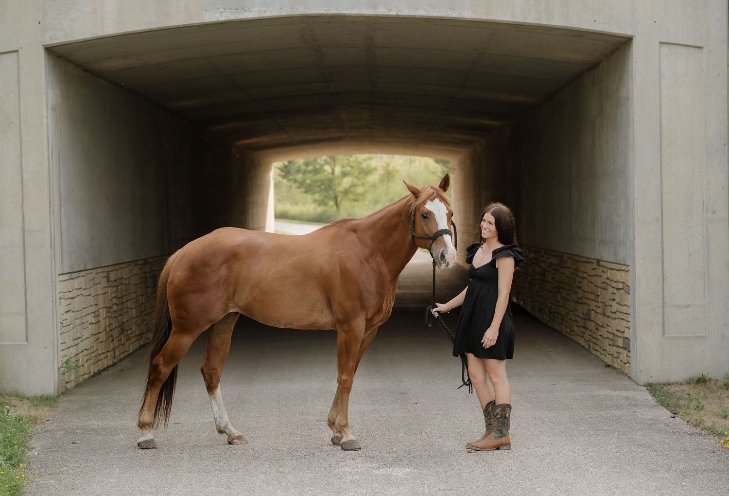 A woman in a black dress and boots stands with a chestnut horse under a concrete underpass.
