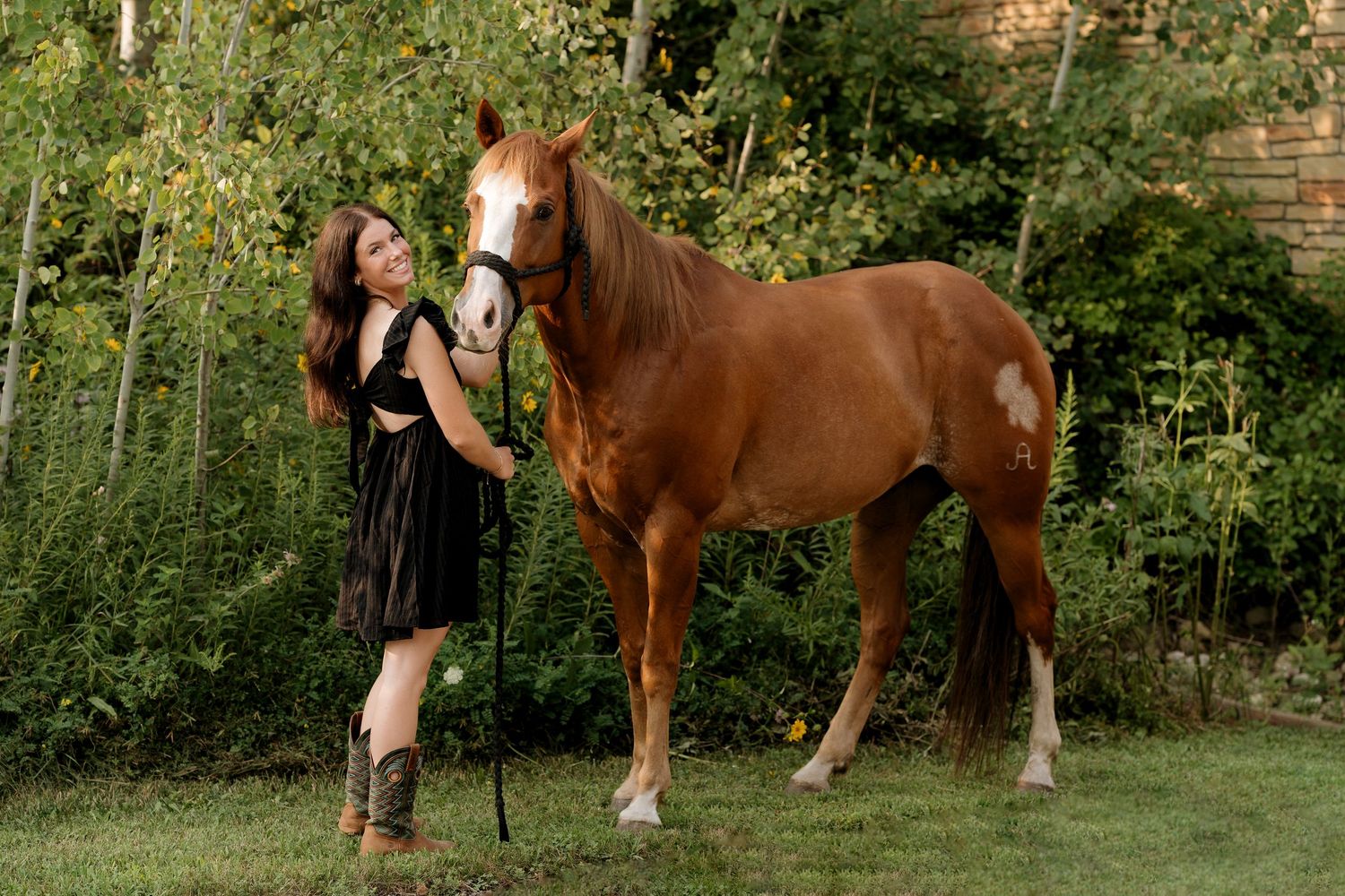 Young woman in black dress and cowboy boots stands beside a chestnut horse on green grass with trees behind.