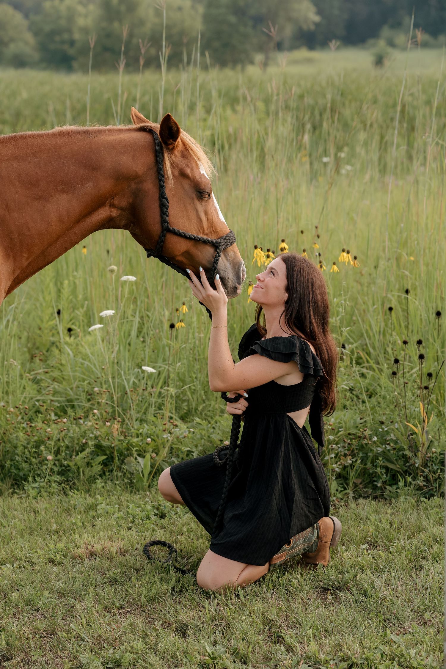Woman in black dress kneeling in field, gently touching nose of chestnut horse with wildflowers in background.