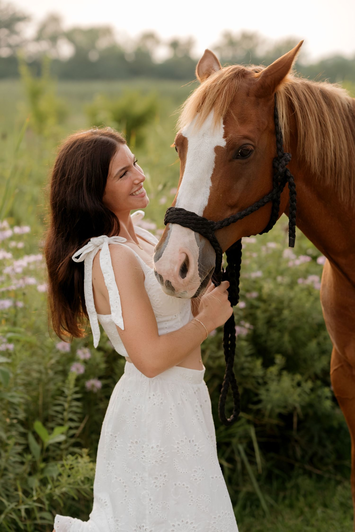 A smiling woman in a white dress tenderly holds a chestnut horse with a blaze in a lush green field.