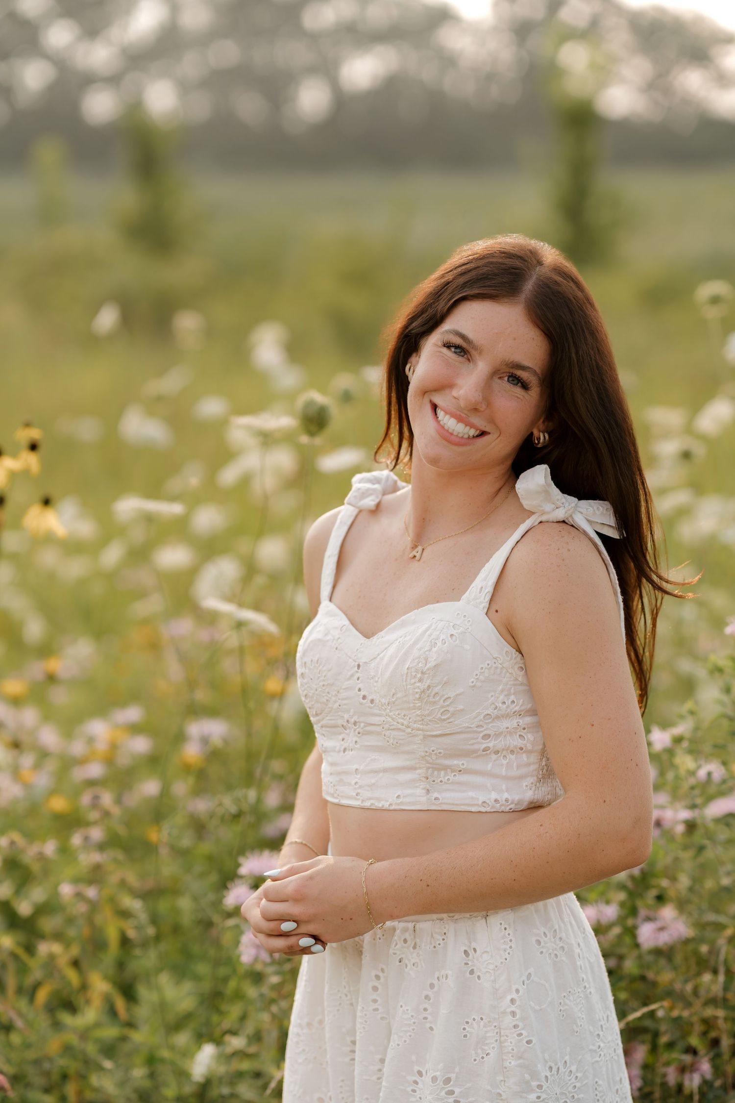 Young woman in white eyelet dress smiling in a wildflower meadow during golden hour.