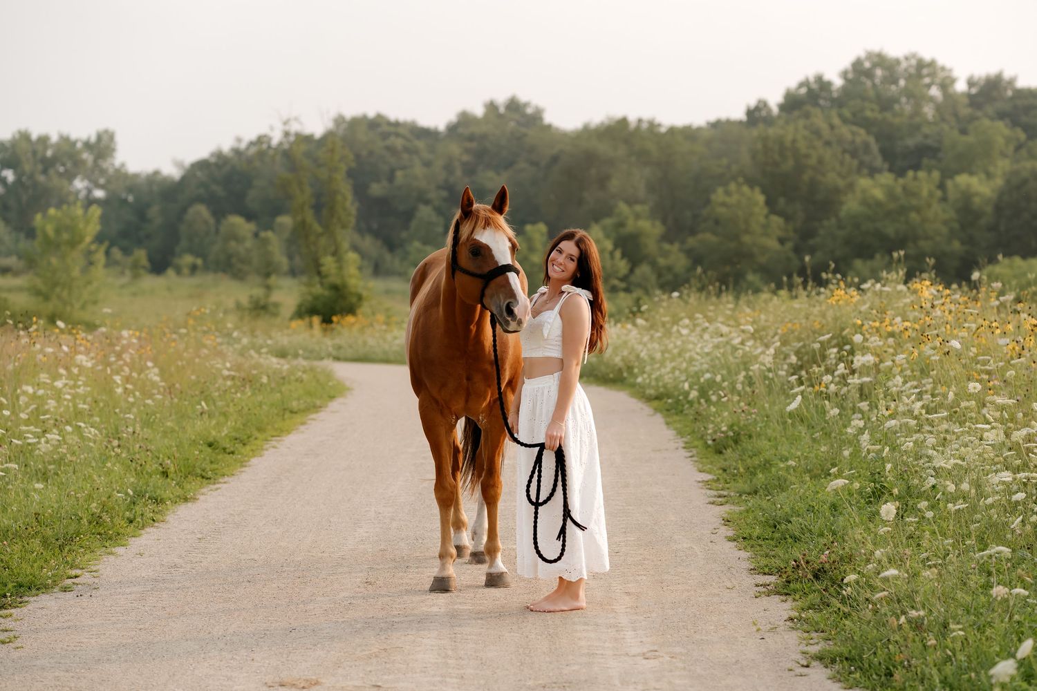 Woman in white dress standing barefoot on a rural path with a chestnut horse amid wildflowers.