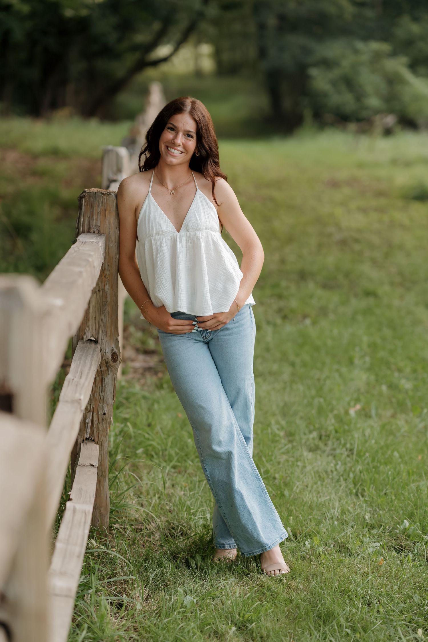 Young woman in white halter top and flare jeans smiling outdoors leaning against a rustic wooden fence.