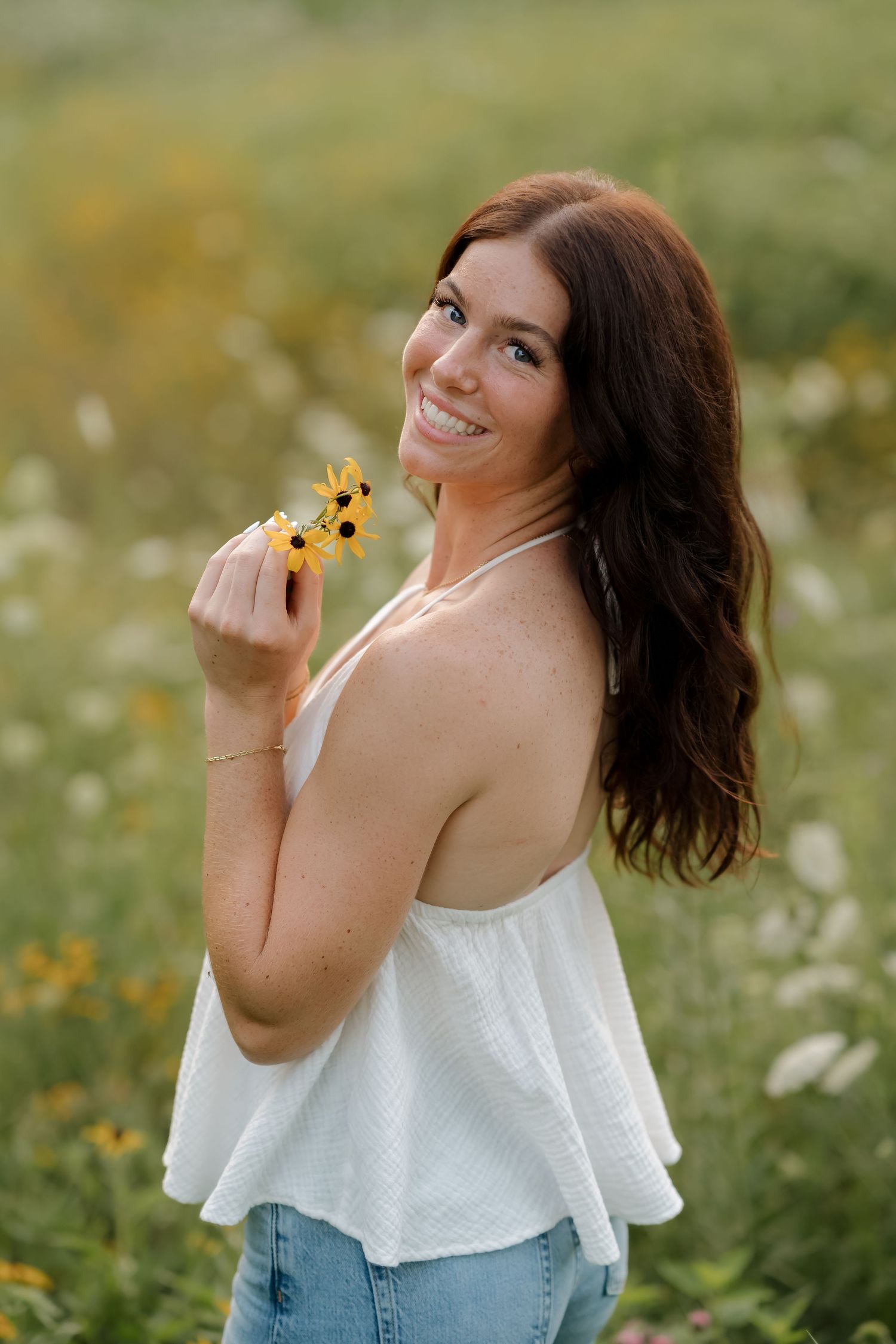Smiling brunette woman holding yellow wildflowers in a white halter top and jeans in a blooming meadow.