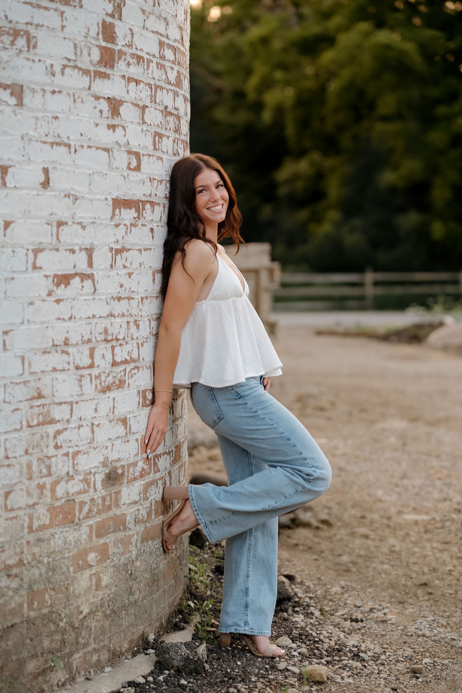 Young woman in white top and flared jeans leaning against rustic brick wall outdoors, smiling confidently.