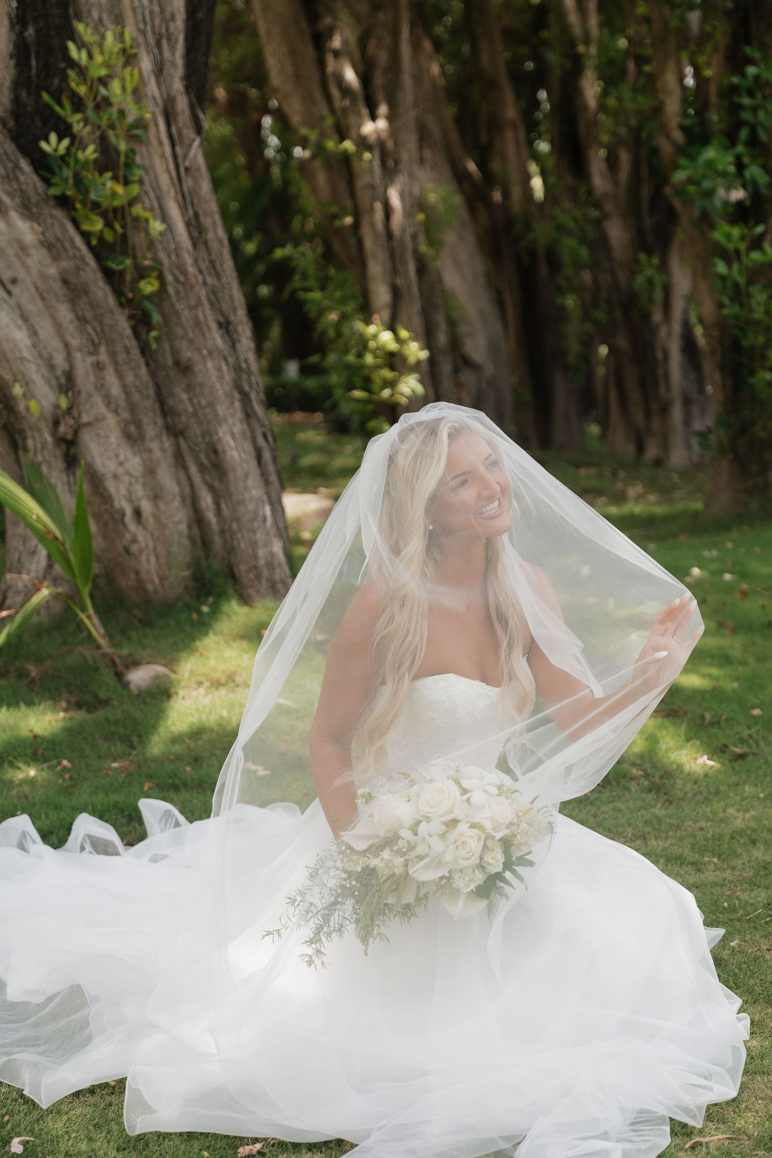 Bride in white gown and veil sits on grass holding bouquet, surrounded by trees in a serene outdoor setting.