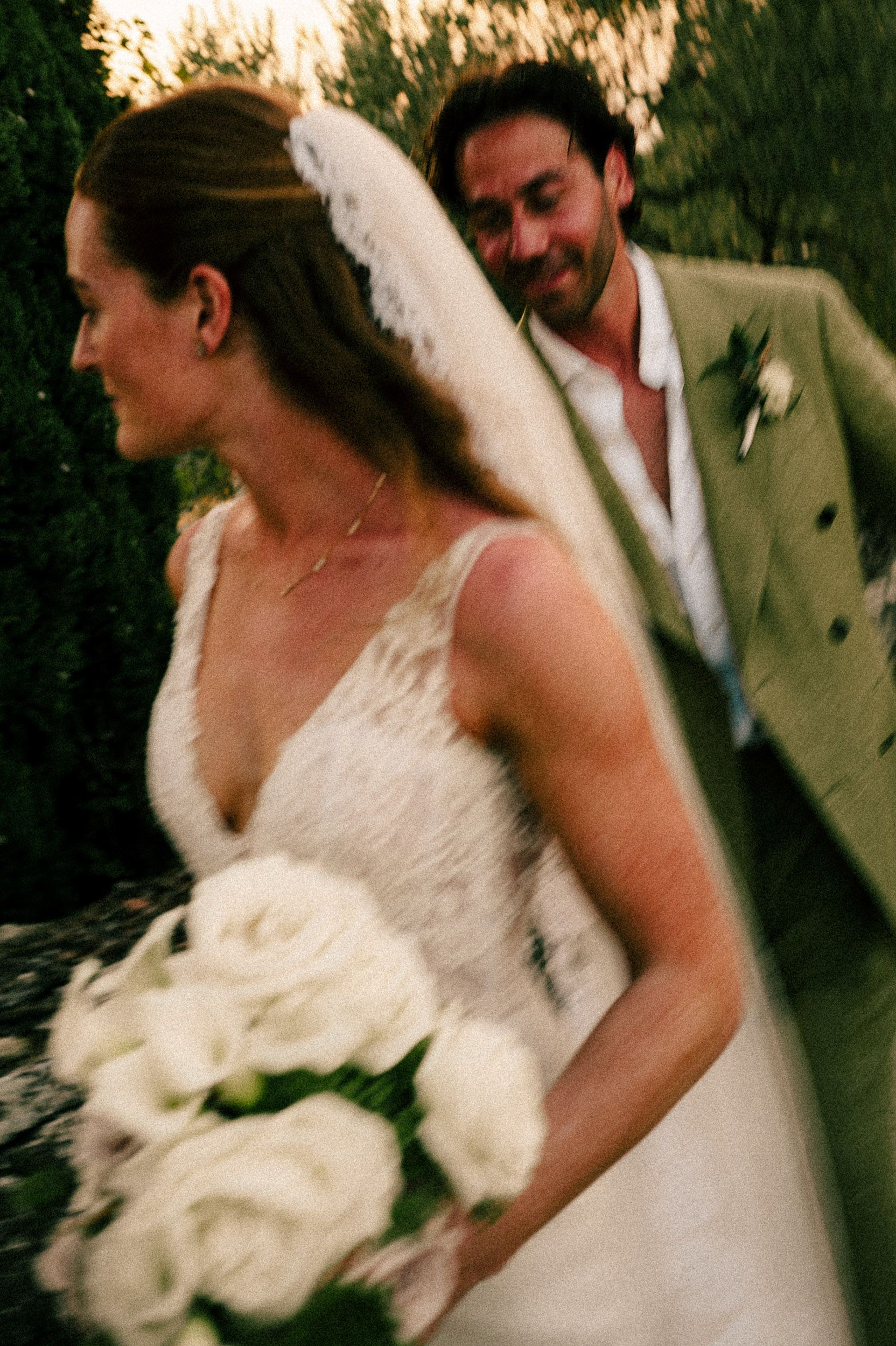 Bride in white lace gown holding white rose bouquet with groom in tan suit smiling behind her at outdoor wedding.