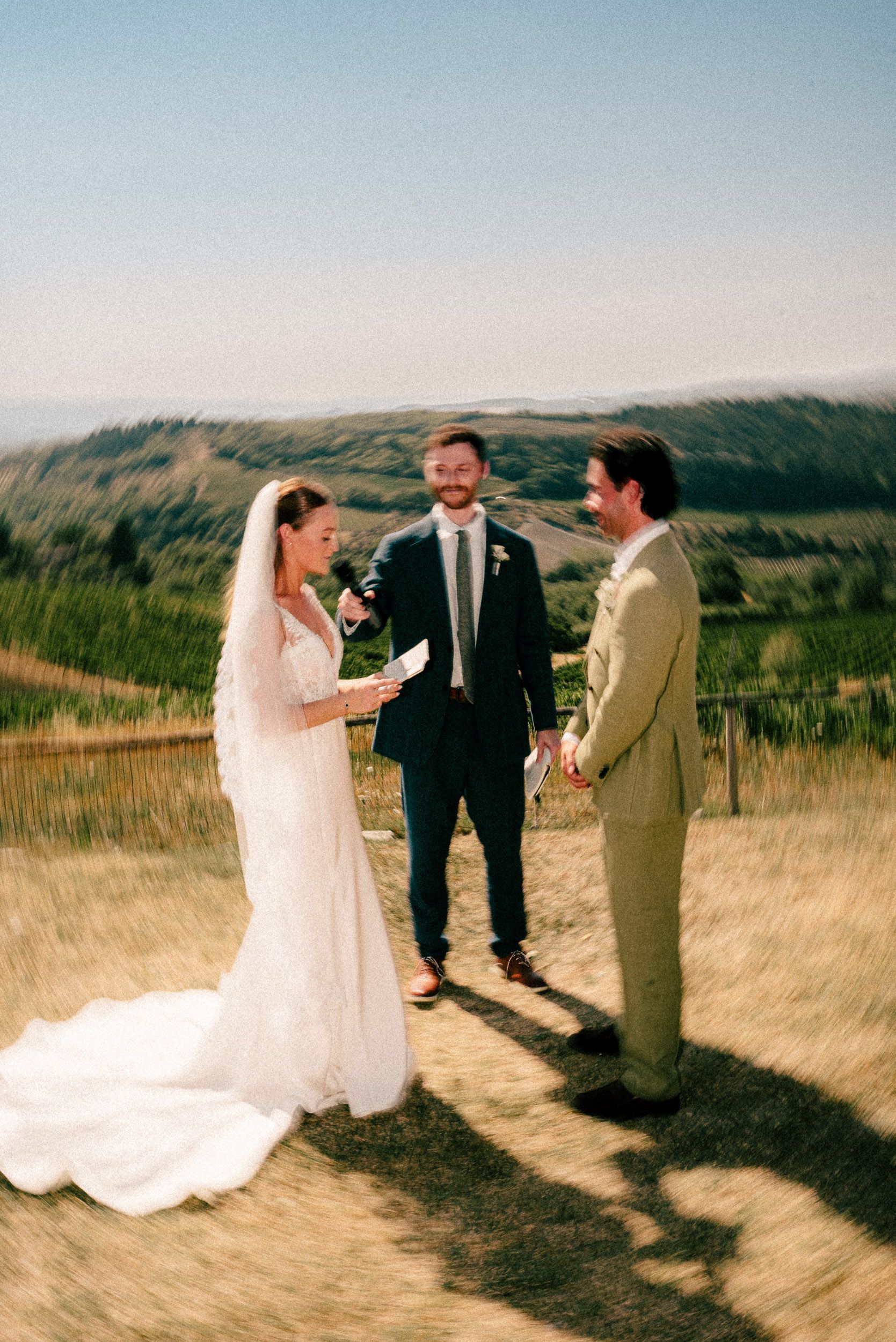 Bride reads vows to groom with officiant at outdoor vineyard wedding ceremony on sunny hillside.