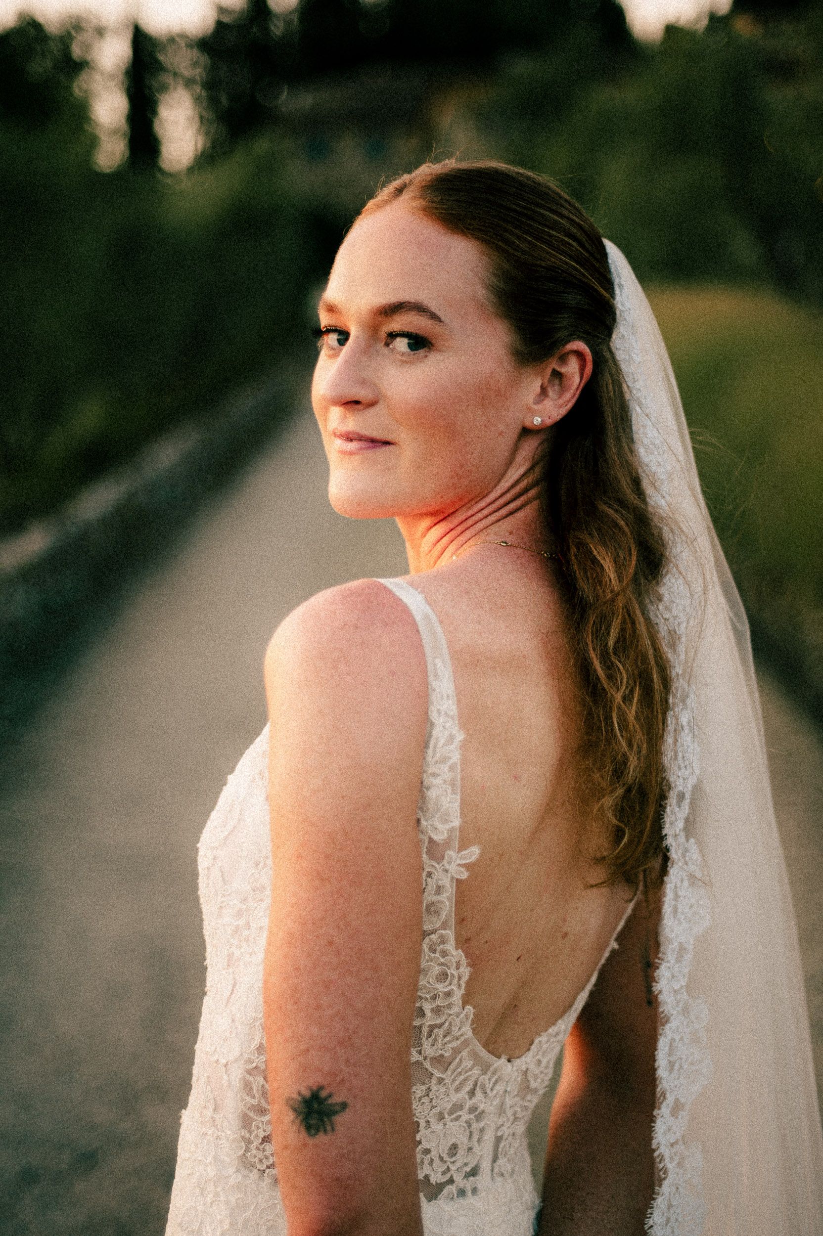 Bride in lace backless wedding dress and veil looks over shoulder at sunset on a tree-lined path.