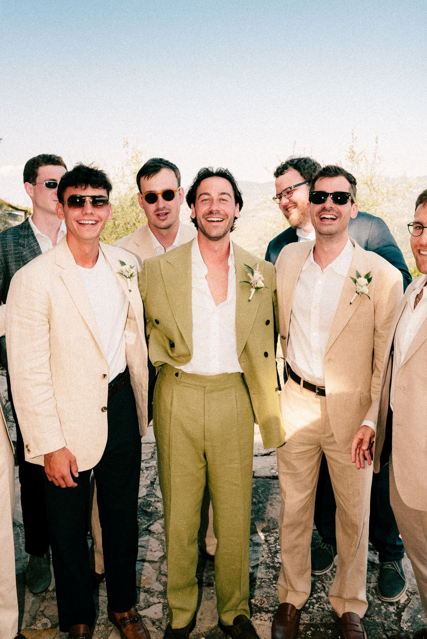 Group of groomsmen in beige suits and sunglasses smiling outdoors at a sunny wedding celebration.