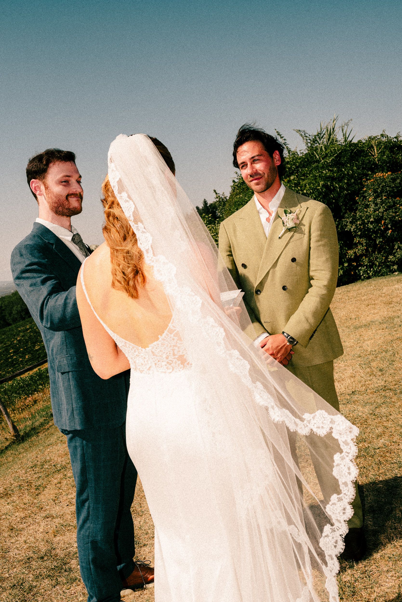Bride in lace veil stands at outdoor wedding ceremony with groom in olive suit and officiant in navy.