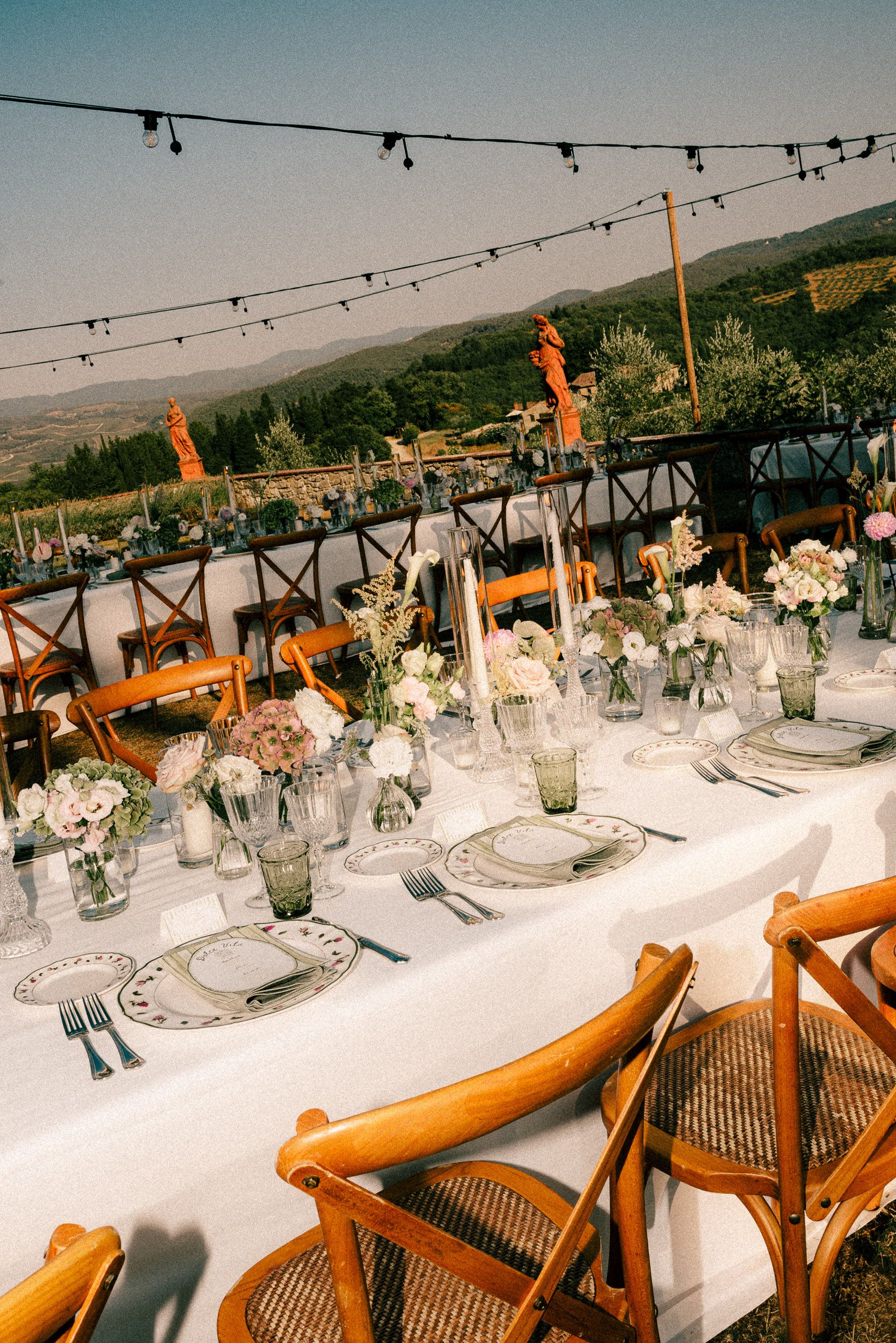 Elegant outdoor wedding reception table with floral centerpieces, string lights, and scenic Tuscan hillside views.