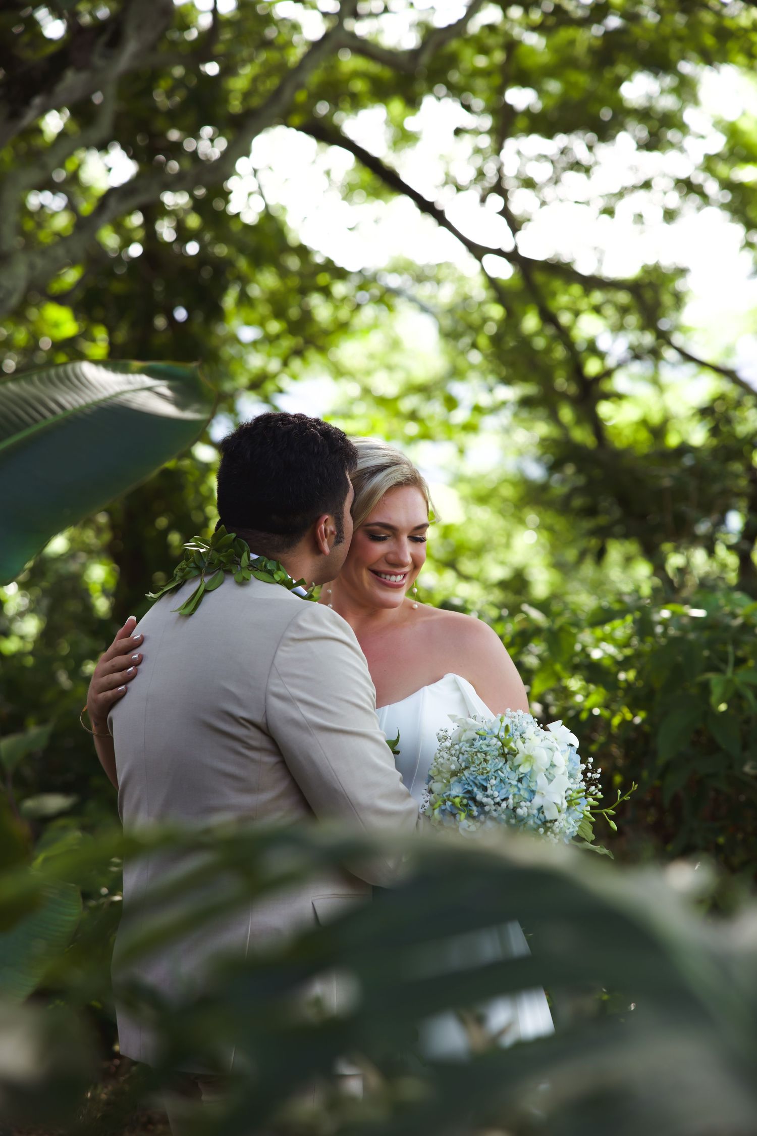 Bride and groom embrace in lush tropical garden, bride holding blue hydrangea bouquet, smiling joyfully.