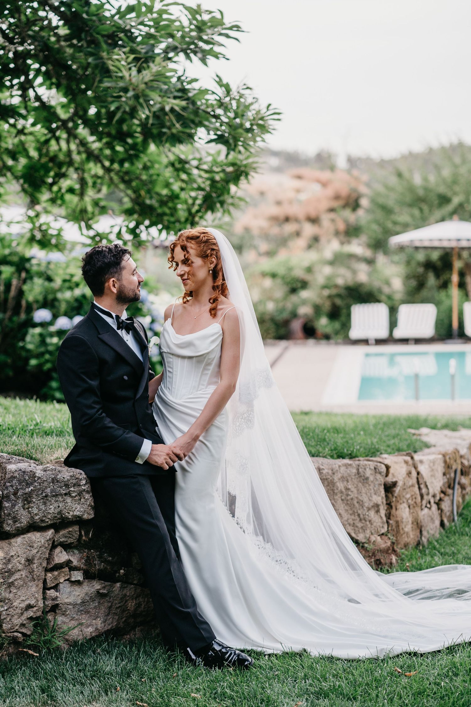 Bride in white gown with veil and groom in black tuxedo share a romantic moment by a stone wall near a pool.