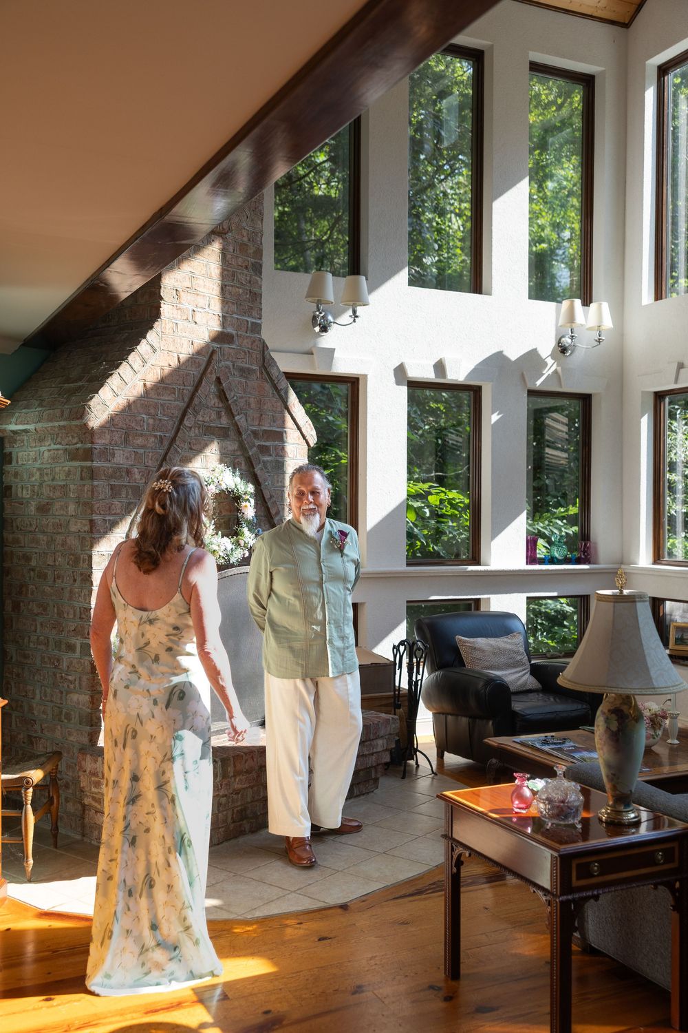 Elderly man in linen suit greeting a woman in floral dress inside a sunlit home with stone fireplace.