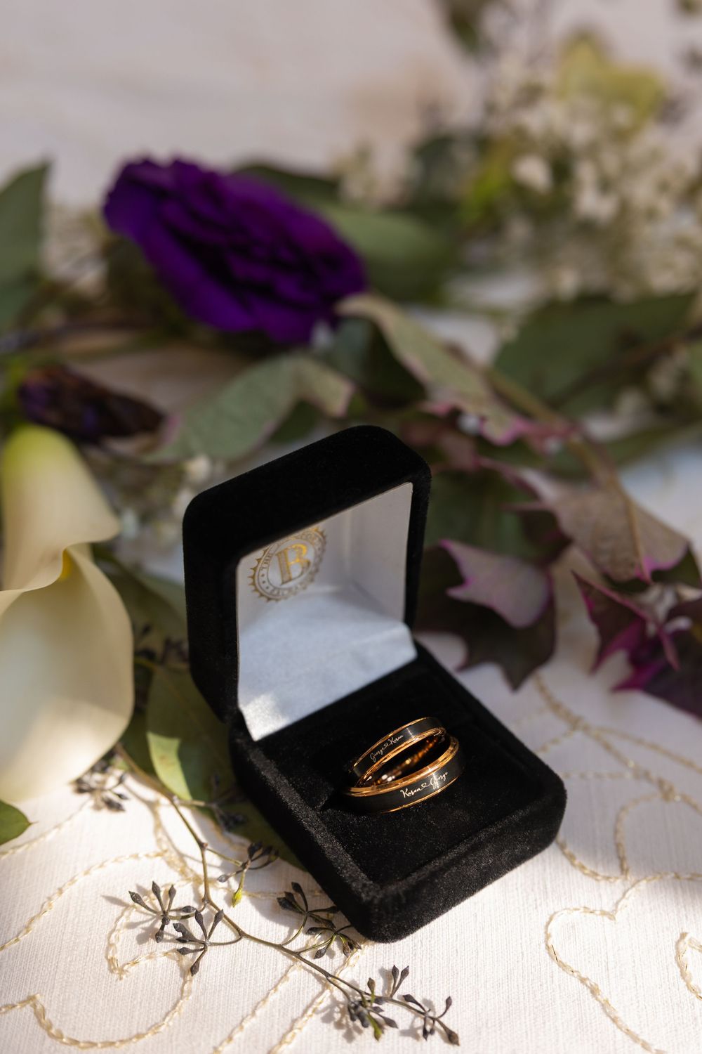 Two gold wedding rings displayed in a black velvet jewelry box surrounded by purple and white flowers.