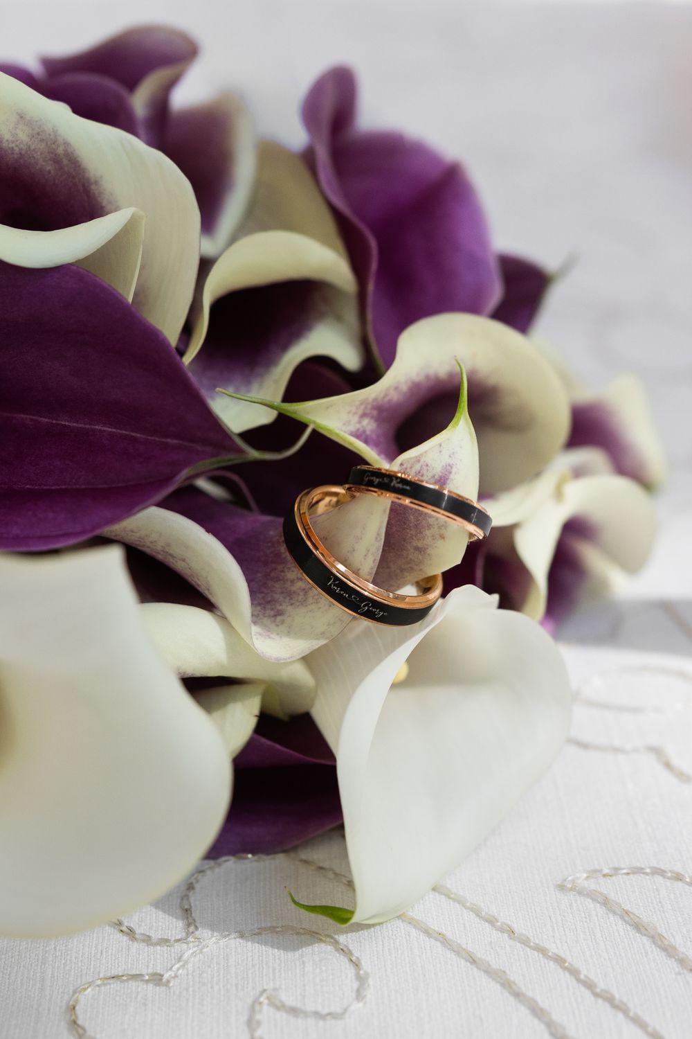 Wedding rings resting on a bouquet of white and purple calla lilies on a white lace surface.