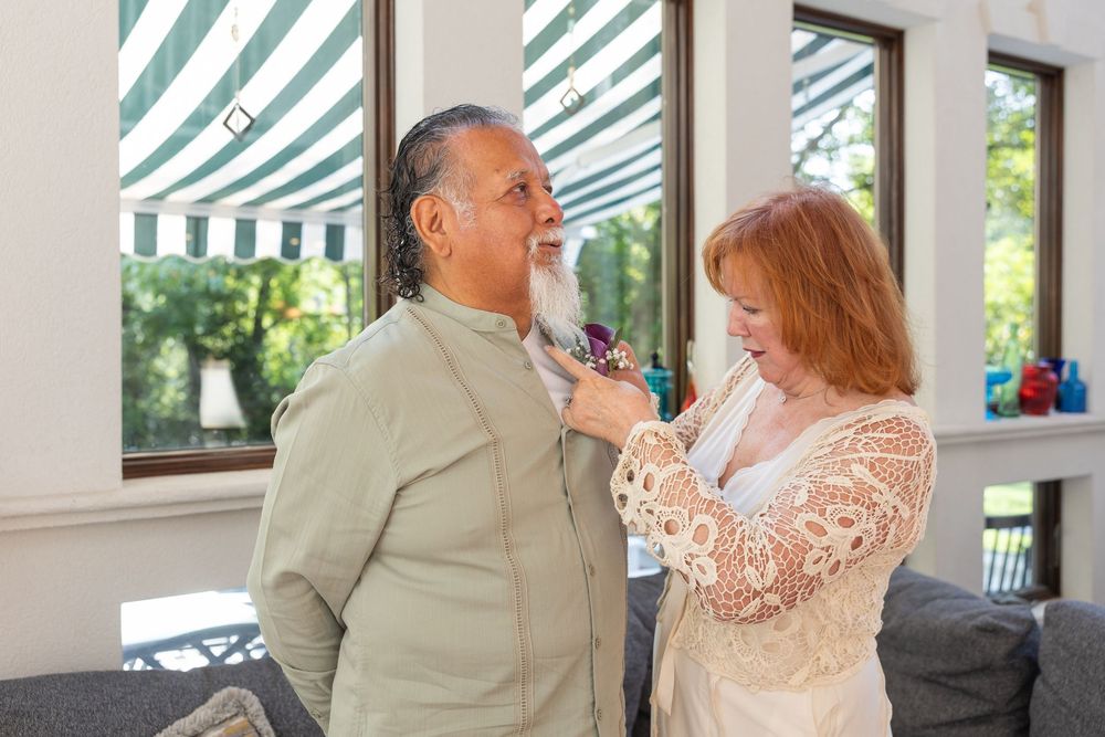 A woman adjusts a man's tie in a bright sunlit room, both dressed elegantly for a special occasion.
