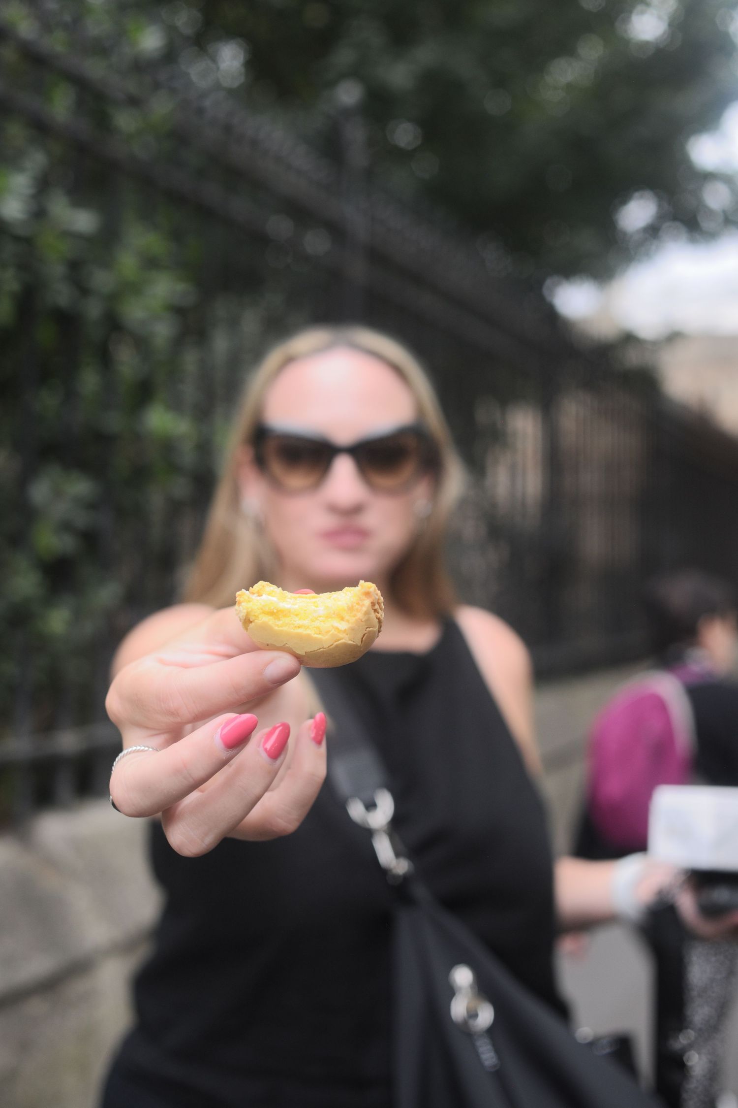 Woman in sunglasses holding a small pastry toward camera outdoors near iron fence.