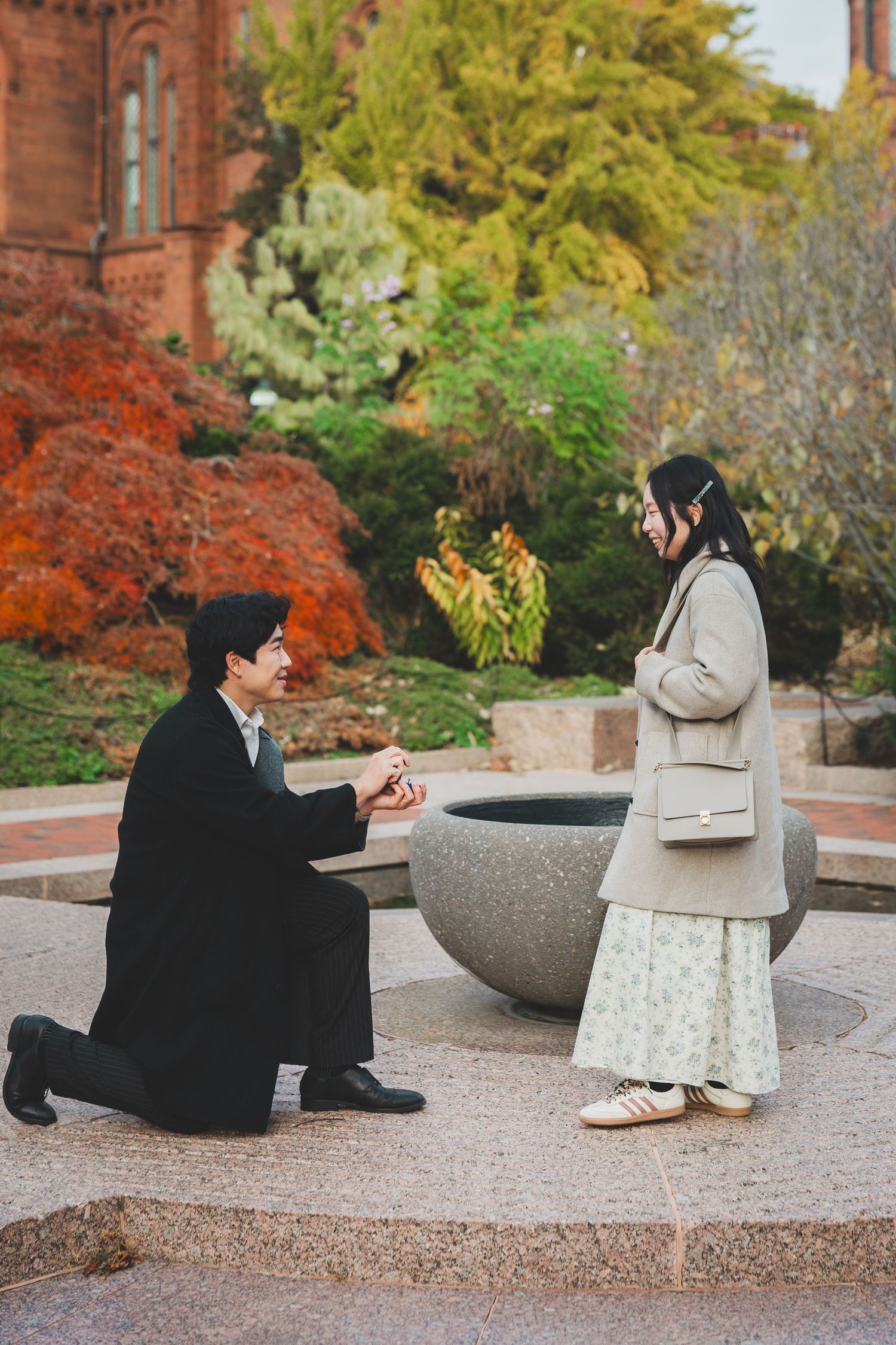 Man kneeling on one knee proposing to a woman in an autumn garden near a stone fountain.