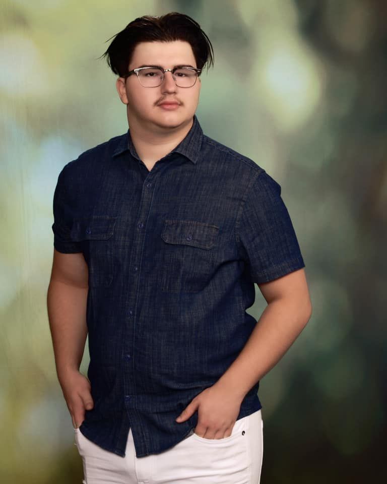 Young man in dark denim shirt and white pants posing for a portrait photo against a green bokeh background.