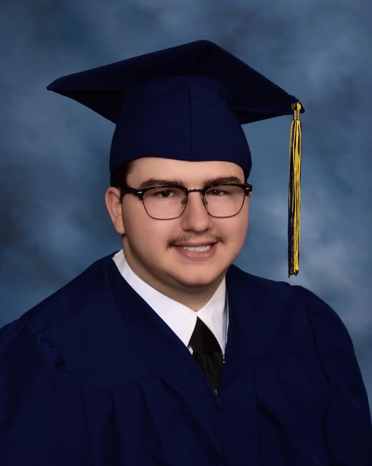 Young man in navy blue graduation cap and gown with gold tassel, wearing glasses, smiling in formal portrait.